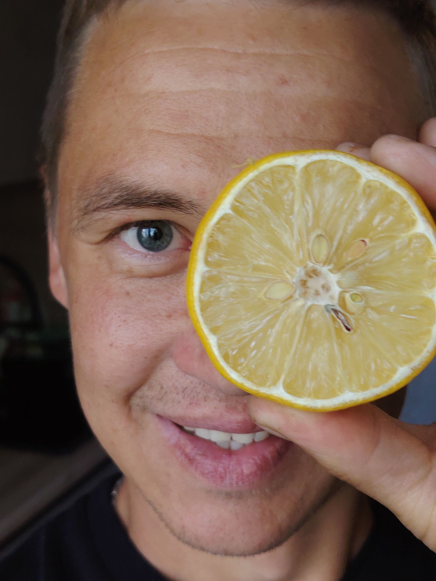 A close-up shot focuses on a person's face, with a slice of lemon held up to cover their right eye. The individual has fair skin, light brown eyebrows, and a grey-blue eye visible. They are smiling, revealing white teeth and pink lips. The background is dark and indistinct, offering no contextual clues about a specific city or location. There is no information in the image to suggest it was taken in Chaikovsky, Russia, or any other urban environment.