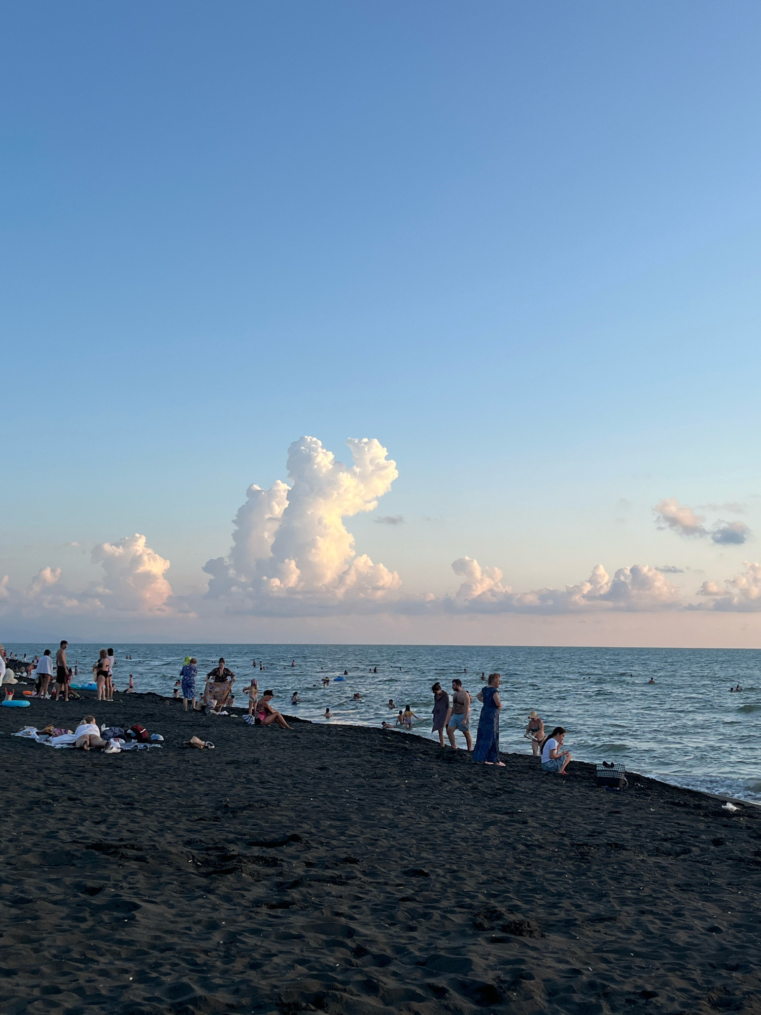 This photograph captures a scene on the black sand beach of Ureki, Georgia, during what appears to be late afternoon or early evening. The sky is a clear, pale blue, transitioning to a softer hue near the horizon where a few scattered, illuminated clouds are present. The sea is a moderate blue-gray with gentle waves lapping at the shore.
On the dark sand beach, numerous people are engaged in various activities. Some are standing and observing the sea, while others are seated or lying down on towels. There are groups of people conversing and children playing. Several individuals are in the water, swimming or wading.
Prominently featured in the background are large, cumulonimbus clouds that have taken on unusual shapes, one resembling a large, anthropomorphic figure and another a pair of connected cloud formations. These clouds are brightly lit from the side, suggesting the sun is low in the sky. The overall ambiance is one of leisure and relaxation on a summer day by the Black Sea.