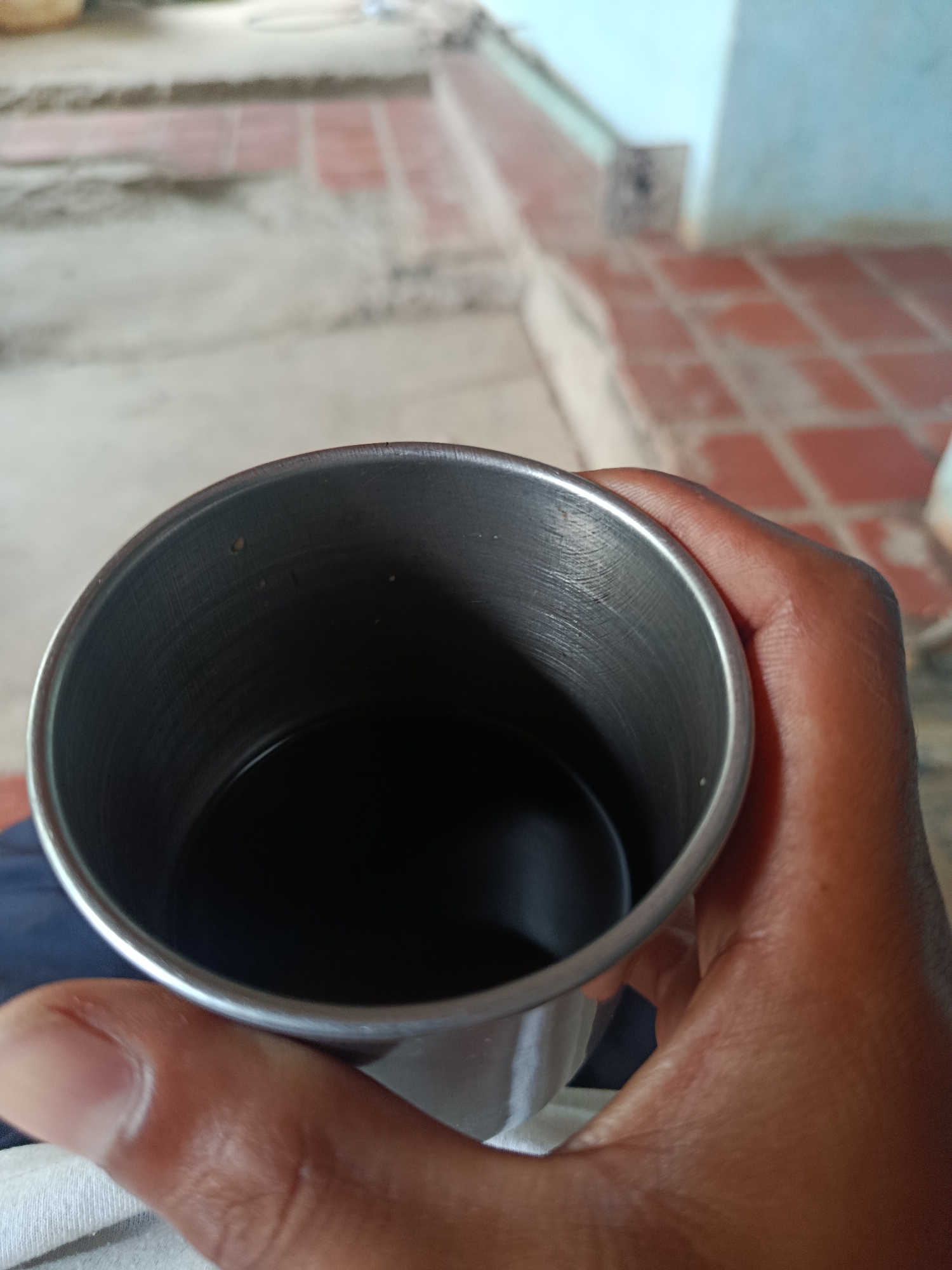 A hand, appearing to be that of a person with darker skin, holds a metal cup filled with a dark liquid. The cup has a brushed metal finish and visible horizontal striations on its interior. The background is blurred, but suggests an outdoor or semi-outdoor setting with tiled flooring and a wall. The lighting indicates it is daytime. The scene does not contain any specific identifiable landmarks of Juan Griego, Venezuela.