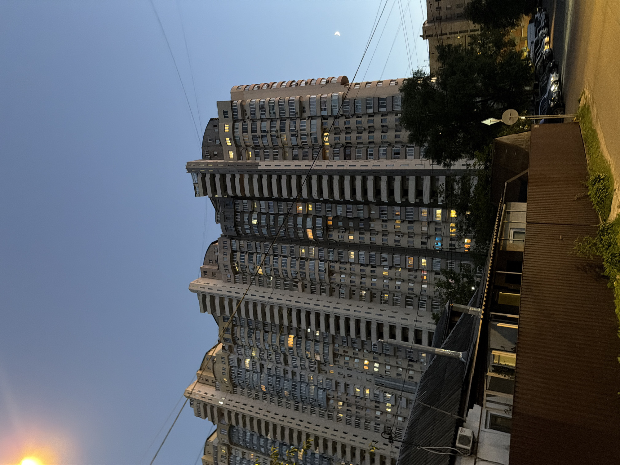 The image shows a tall, multi-story apartment building in Kyiv, Ukraine, during the evening. The building features numerous balconies and windows, many of which are lit, suggesting inhabitants are present. In the foreground, there's a lower building with a corrugated metal roof and a fence. A street with parked cars is visible to the right, and a street lamp is illuminated in the upper left corner. The sky is a clear, deep blue with a crescent moon visible in the upper right quadrant. The overall impression is of a residential urban scene at dusk.