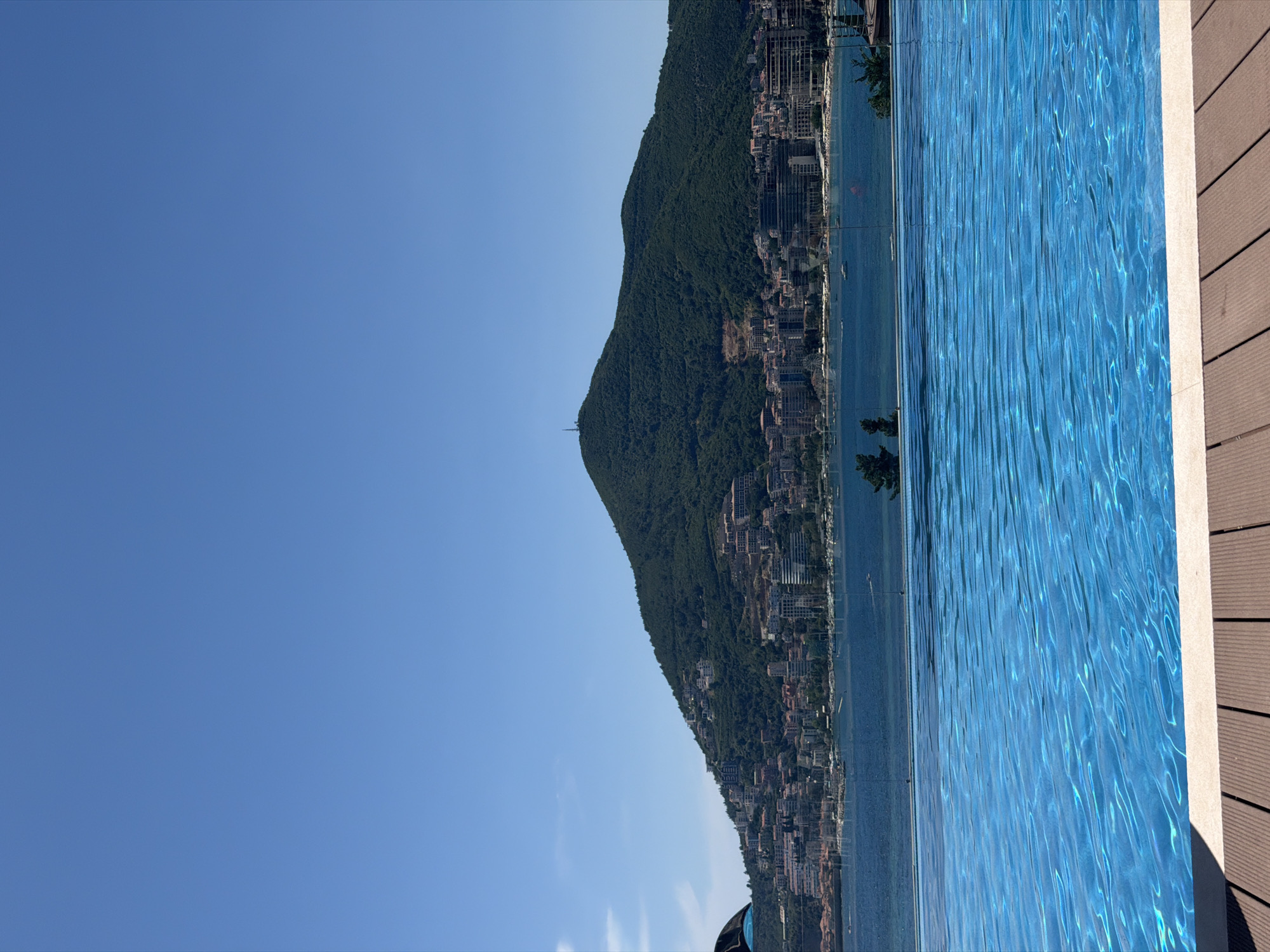 The image depicts a scenic view of the city of Budva, Montenegro, from a vantage point overlooking the Adriatic Sea. In the foreground, a clear blue swimming pool with rippling water occupies the lower half of the frame, bordered by a wooden deck. A glass railing separates the pool area from the bay.
Beyond the pool, the deep blue sea stretches to the horizon. Across the water, the city of Budva is nestled along the coastline, with buildings cascading up the verdant slopes of a large, tree-covered mountain that dominates the background. A prominent telecommunications tower is visible atop the mountain. The sky is a clear, bright blue, indicating a sunny day. The scene conveys a sense of tranquility and leisure, characteristic of a resort destination.