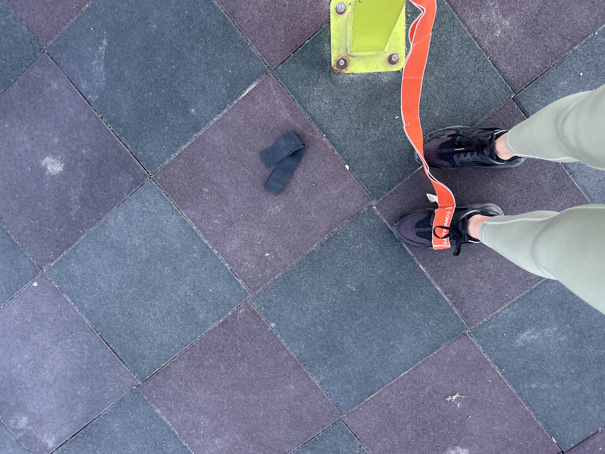 The image shows an overhead view of an outdoor workout area, identifiable by the interlocking rubber floor tiles in alternating dark blue and purple hues. A person wearing light green leggings and black sneakers stands near a green metal fitness structure; an orange resistance band is attached to the structure, looping partially around the left sneaker. A black resistance band lies loose on the rubber floor tiles to the left. The location is in Burgas, Bulgaria.
