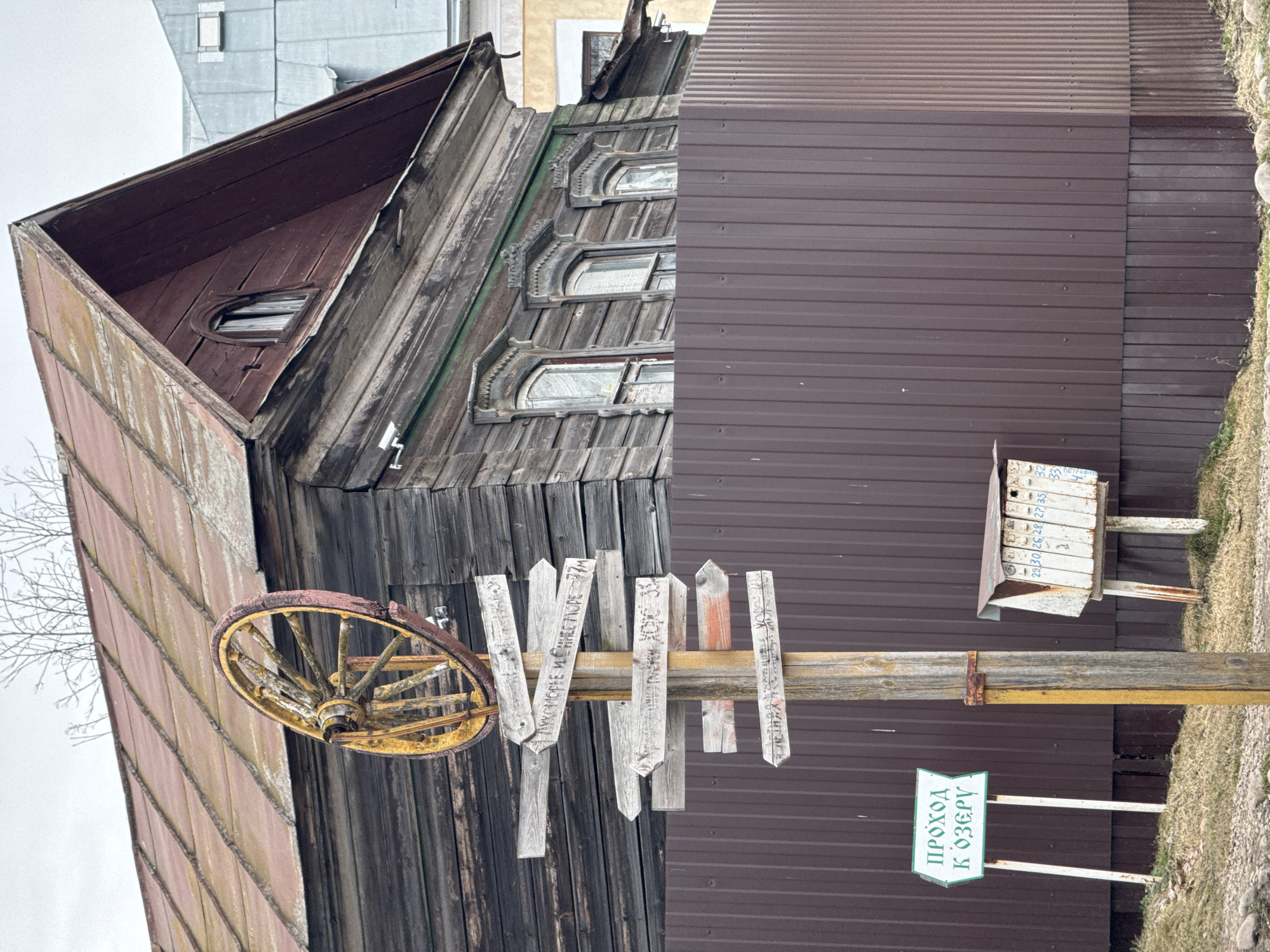 The image depicts a rustic scene in Kladovitsy, Russia, showcasing a weathered wooden building with a combination of traditional and modern elements. The building's façade is partially clad in brown corrugated metal siding, contrasting with the older, dark, and aged wooden planks forming the original structure. A distinctive feature of the wooden section is the presence of three ornate, arched windows with decorative trim.
Attached to the building is a yellow, rusted wagon wheel, suggesting a decorative or symbolic purpose. A wooden signpost stands in front of the building, displaying several weathered, hand-painted directional signs, some of which appear illegible. A sign near the bottom of the post reads "ПРОХОД К ОЗЁРУ" (Passage to the Lake). A mailbox sits on the right side of the signpost. The building's roof features an oval window. The overall setting is likely a rural or semi-rural area, with visible vegetation and a overcast sky.