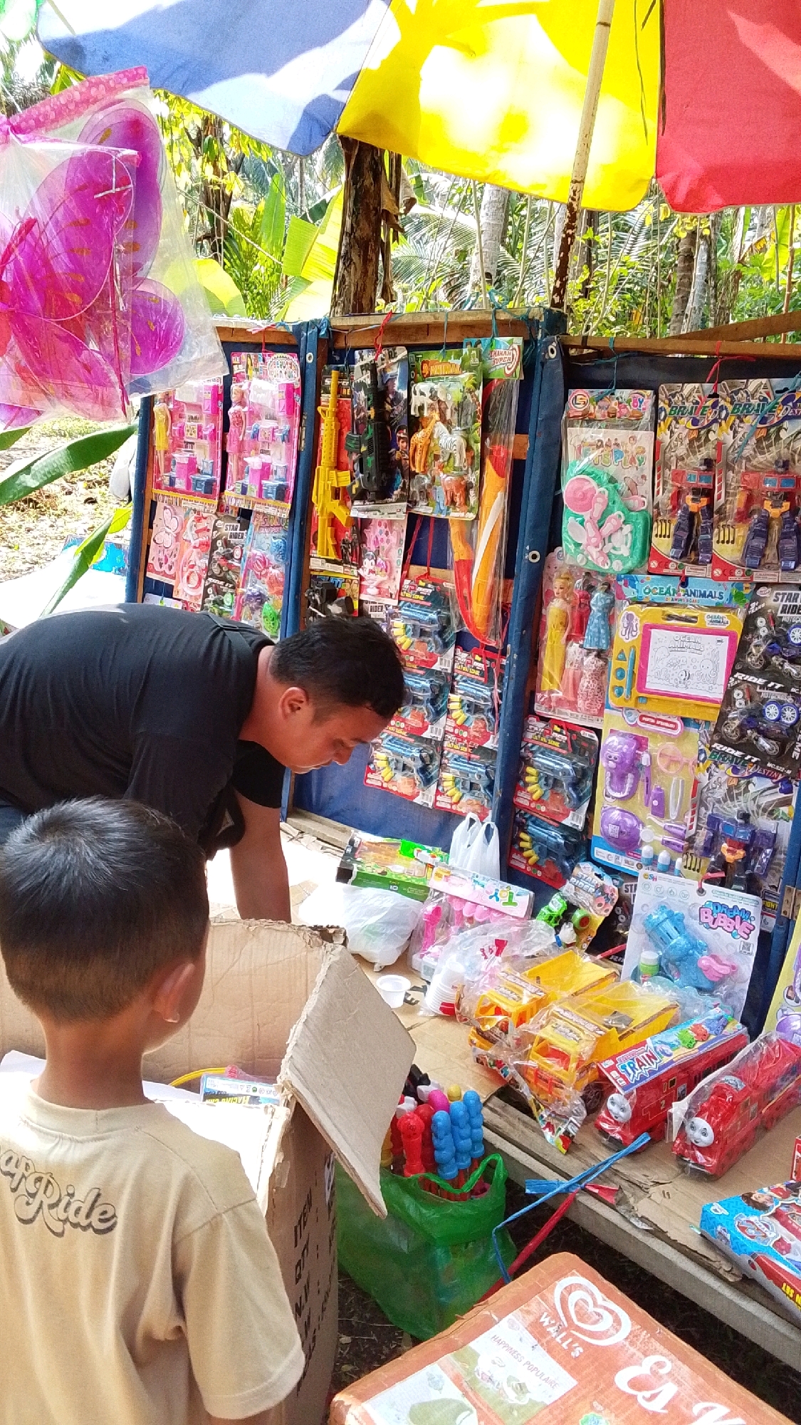 In Parakanmanggu, Indonesia, a man in a black t-shirt is bent over a stall displaying a variety of toys. A young boy, seen from the back wearing a tan t-shirt with the word "Ride" printed on it, stands near a cardboard box. The stall is covered by colorful tarpaulins in blue, yellow, and red. The toys include toy guns, action figures, toy animals like zebras and elephants, and toy vehicles such as trains and trucks. Some toys are packaged, while others are loose. Several plastic bags are visible, and a green plastic bag holds a bunch of colorful items that appear to be toy whistles or similar small toys. The ground appears to be dirt and leaves. The overall scene depicts a temporary outdoor toy vendor.