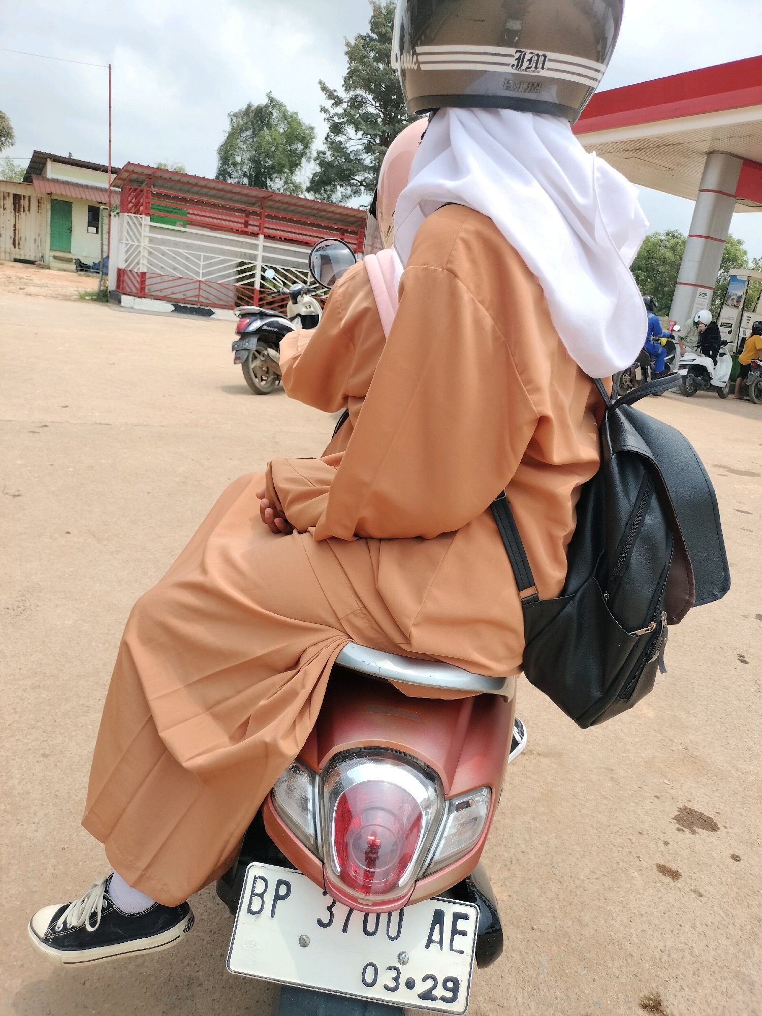 Two individuals are seated on a scooter, with the foreground person wearing a tan abaya and black Converse sneakers, and a black backpack. The scooter is parked in front of a gas station, likely on a street in Batam, Indonesia, given the license plate's format which includes "BP" and "AE", commonly seen in Riau Islands Province. The environment appears to be a warm, outdoor setting with a paved surface, a building with a red-trimmed roof in the background, and trees. The sky is overcast.