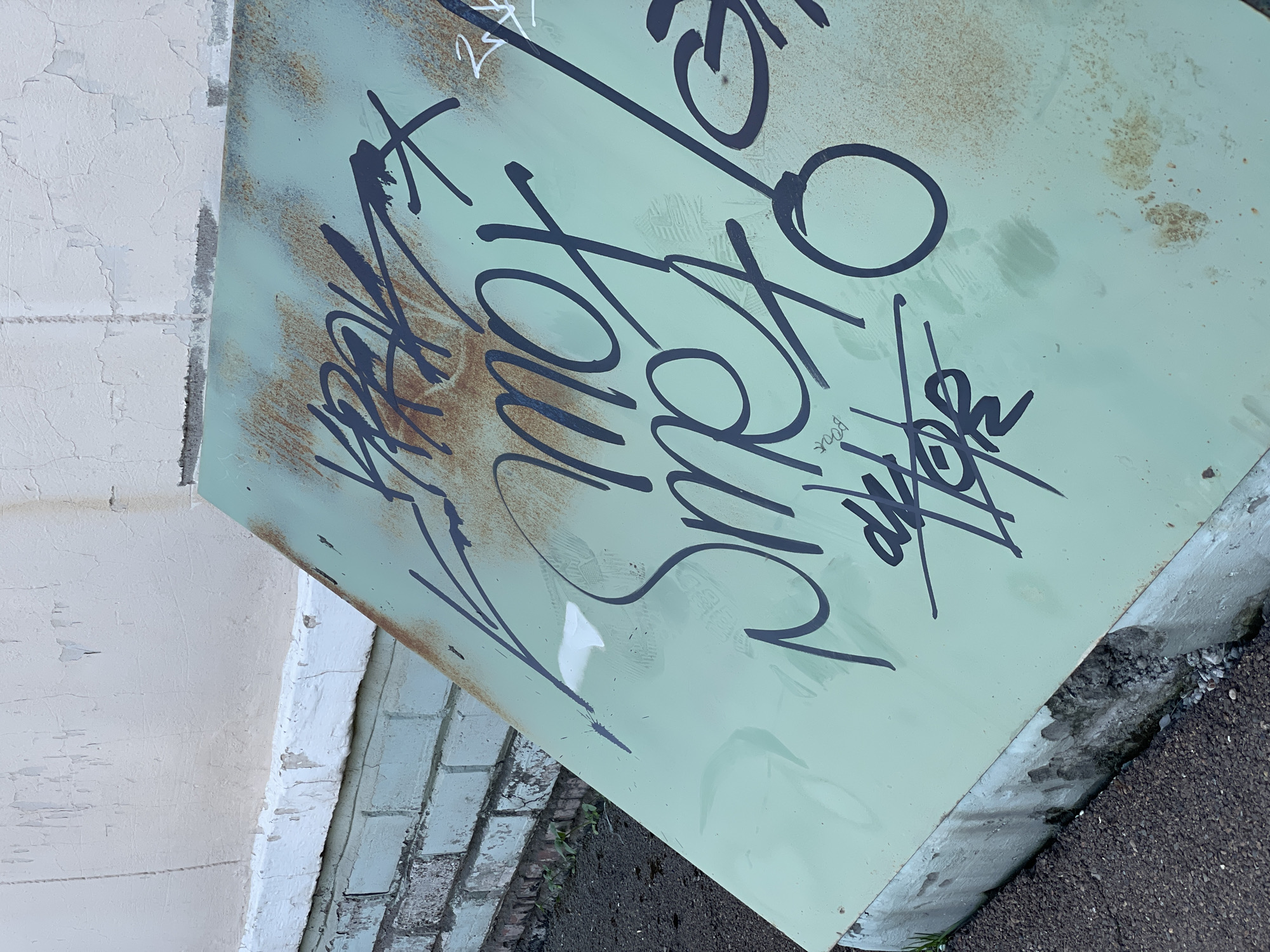 A tilted, rectangular, light green metal surface with rust spots and black graffiti leans against a white brick wall and a grey concrete structure. The graffiti consists of stylized lettering and tag-like symbols. The surrounding environment includes cracked white paint on a wall, aged brick, and asphalt. The image does not provide any information indicating it is in Zheleznogorsk, Russia.