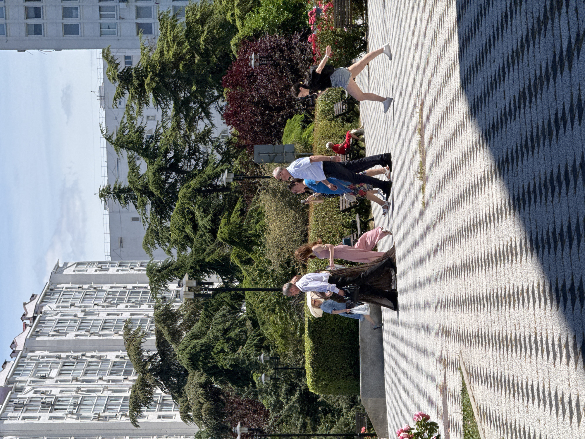 In a city park in Unknown, Unknown, a group of people are walking on a patterned pavement. Several tall, modern apartment buildings are visible in the background, with lush green trees and shrubs in between. The people, dressed in summer attire, are walking in various directions. One woman in a black top and denim shorts is running or in motion, with her arms outstretched. Another woman in a long, flowing pink dress is walking behind her. A couple, a man in a blue striped shirt and dark pants and a woman in a light blue dress carrying an umbrella, are walking together. A person in a red outfit is seated on a bench in the distance, and several other individuals are blurred in the background. The sun casts strong shadows on the patterned walkway, suggesting a bright, sunny day. The park appears to be well-maintained, with blooming pink roses visible in the foreground.