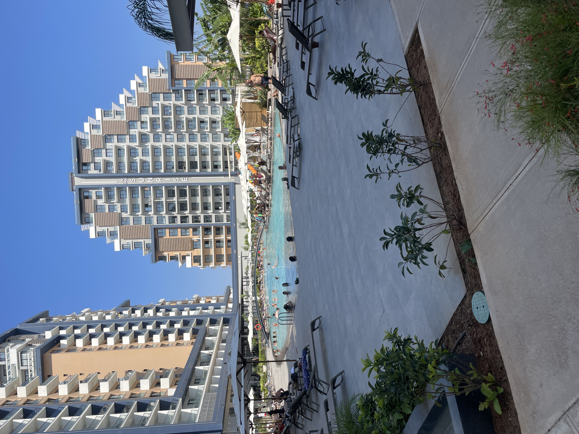 This image depicts a large outdoor swimming pool area at a resort, likely the Merit Crystal Cove Hotel in Trikomo, Northern Cyprus, given the architectural style of the buildings which are visible in the background. Two modern, multi-story buildings with numerous balconies and a distinctive stepped facade flank the pool. The pool itself is filled with people, some swimming, others wading or standing. There are numerous lounge chairs and umbrellas set up around the pool on a paved area. Greenery, including palm trees and smaller plants, is scattered throughout the area. The sky is clear and blue, indicating a sunny day. The overall scene is one of a bustling resort environment, with guests enjoying leisure activities by the pool.