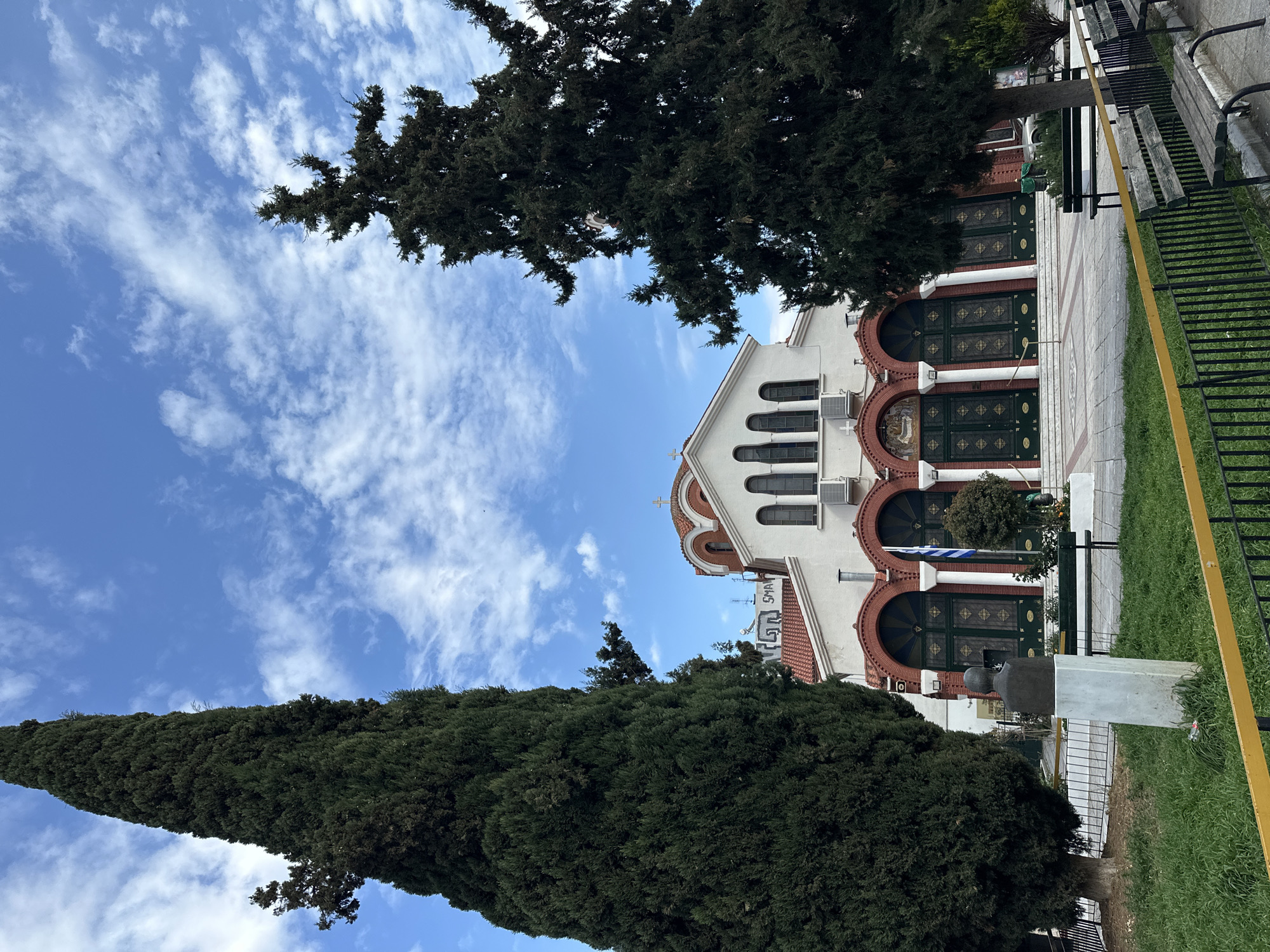 The image depicts a church in Serres, Greece, partially obscured by trees. The church is white with a red-tiled roof and arched doorways. The doorways are dark green with decorative gold accents. A Greek flag is visible near one of the doorways. The sky is blue with scattered white clouds. The trees framing the church are tall and green. A metal fence is on the right, with a yellow bar running along the top. A small statue is visible near the base of one of the trees.