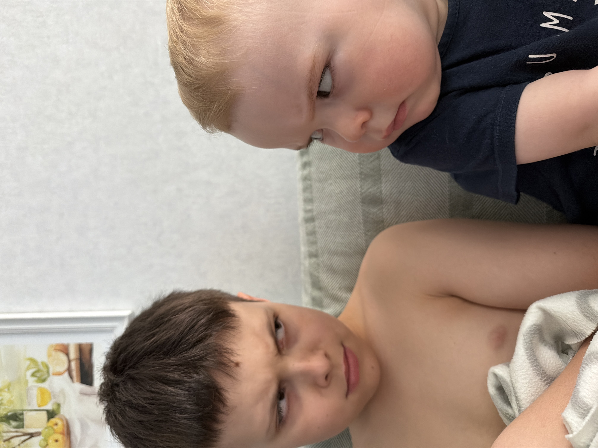 This portrait-oriented image captures an intimate indoor scene featuring two children, likely brothers, lying down on what appears to be a couch or bed in a home in Votkinsk, Russia.
The younger child, positioned towards the top of the frame, has fair skin and light blond hair. He is wearing a dark blue t-shirt with white lettering; partial letters "M" and "W" are visible. He looks intently downwards and to his left, seemingly observing his older sibling, with a focused and slightly serious expression.
The older child, positioned towards the bottom of the frame, has fair skin and short, dark brown hair. He appears to be shirtless or wearing a very light top, with a grey and white striped blanket or towel partially covering his side. He looks towards the viewer with a subtle smirk or thoughtful gaze.
The background consists of a light gray, subtly textured wall on the left, and a neutral-toned, possibly woven fabric (like a cushion or blanket) on the right, providing a soft backdrop. A framed picture, depicting a still life with fruits and possibly bottles, is partially visible behind the older child's head, adding a domestic touch. The overall lighting is soft and even, indicating an indoor setting. The image captures a quiet, personal moment between the two children.