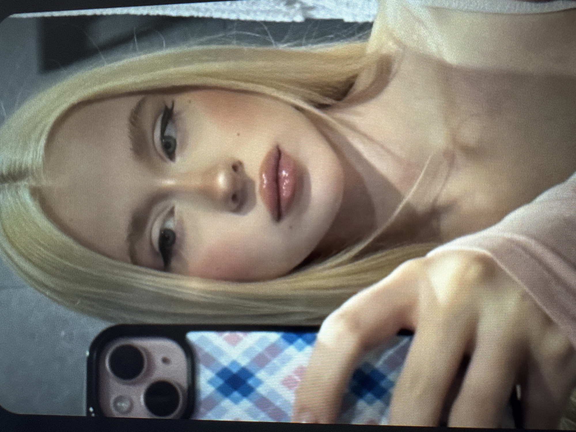 A close-up selfie captures a young person, likely female, with fair skin and long, straight blonde hair, lying down with her head tilted to the left. Her gaze is directed towards the camera with a neutral and serene expression. Her facial features include full, glossy lips, prominent winged eyeliner, and a subtle blush on her cheeks. Her blonde hair is parted down the middle and frames her face.
In the lower right foreground, her right hand is visible, holding a smartphone. The phone's dual camera module is clearly seen, and it is enclosed in a case featuring a distinct blue and white checkered or argyle pattern. Her fingers are loosely wrapped around the device. She appears to be wearing a light-colored, possibly nude or pale pink, soft-textured garment, with her shoulder and upper arm partially visible.
The background is minimalist and out of focus, consisting of a plain, light-toned surface, suggestive of an indoor setting such as a wall or headboard. The lighting is soft and even, highlighting her features. Horizontal lines are visible across the image, indicating that it is likely a photograph taken of a digital screen. There is no visible text in the image.
