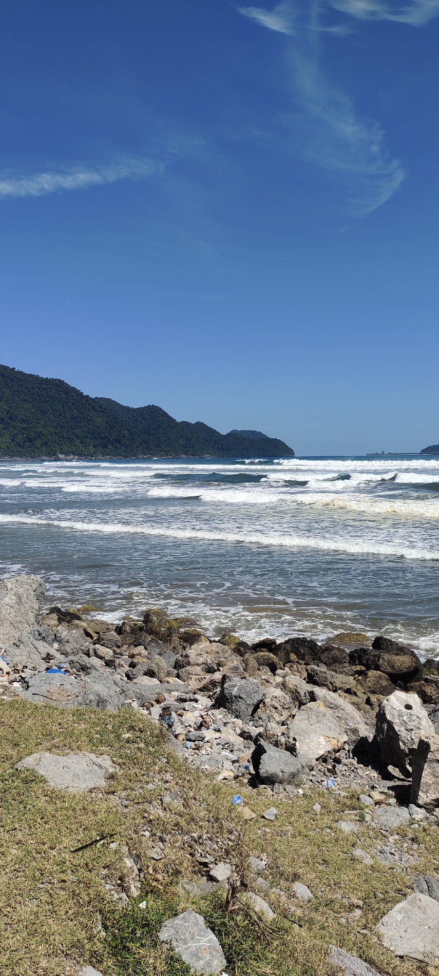 The image shows a coastal scene with a rocky shoreline in the foreground, leading to the ocean with moderate waves. Lush green, tree-covered mountains form a backdrop to the sea. The sky is clear blue with a few wispy clouds. The scene is devoid of people. The rocky area in the foreground shows some scattered debris, including plastic bottles and wrappers, indicating potential environmental concerns. The overall impression is a beautiful natural landscape affected by human presence. The location is identified as Meudheun, Indonesia.