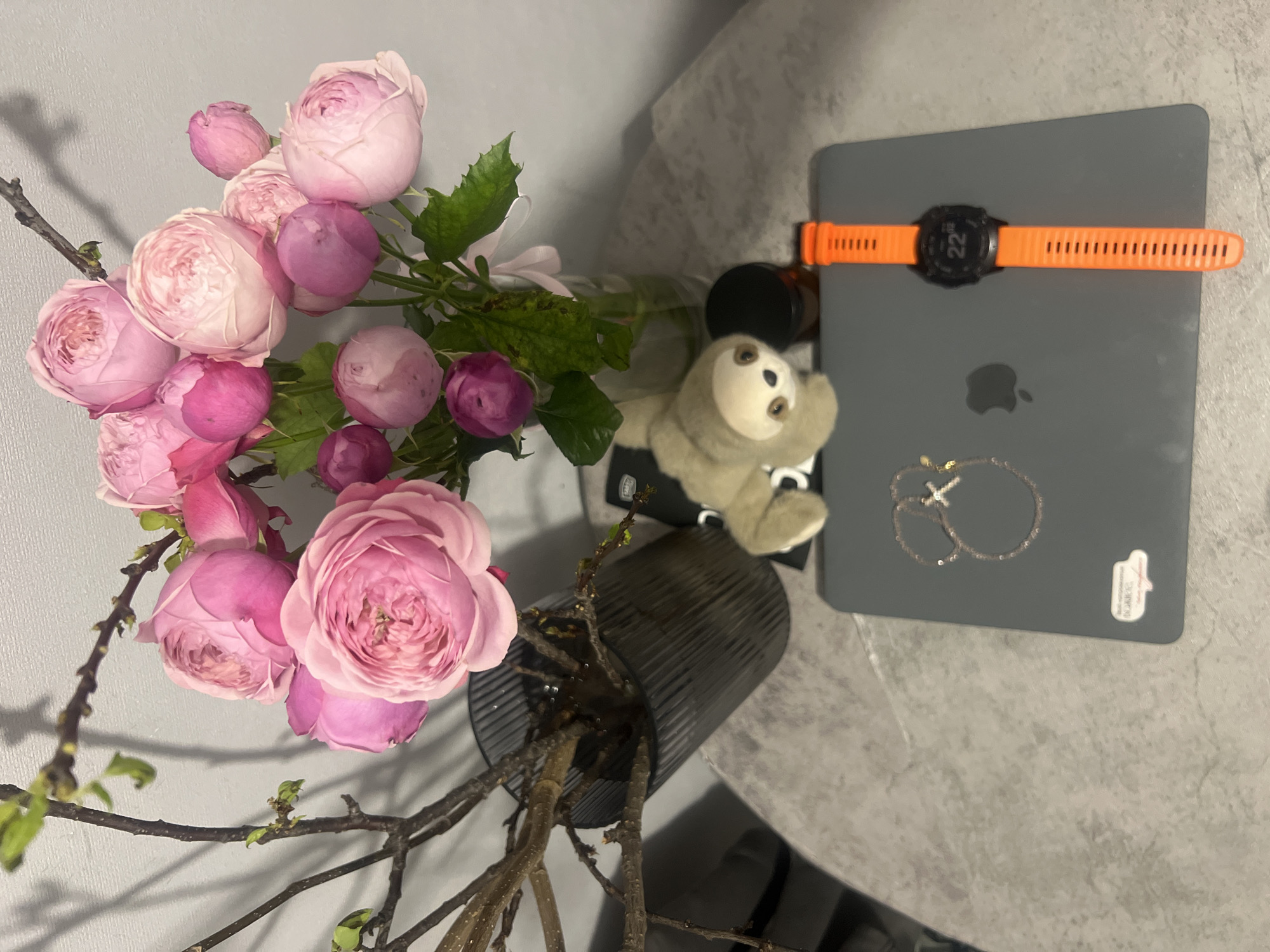 The image depicts a still life arrangement on a gray, marble-patterned surface. A bouquet of pink roses in a dark, fluted vase occupies the left side of the image. To the right of the vase is a gray laptop with an Apple logo. On top of the laptop, there is an orange fitness watch, a brown glass bottle with a black cap, a small sloth plush toy, and a silver-colored necklace. No people are visible in the image. The environment suggests an indoor setting, possibly a home or office. The location is not discernible from the image and cannot be identified as Kazan, Russia.