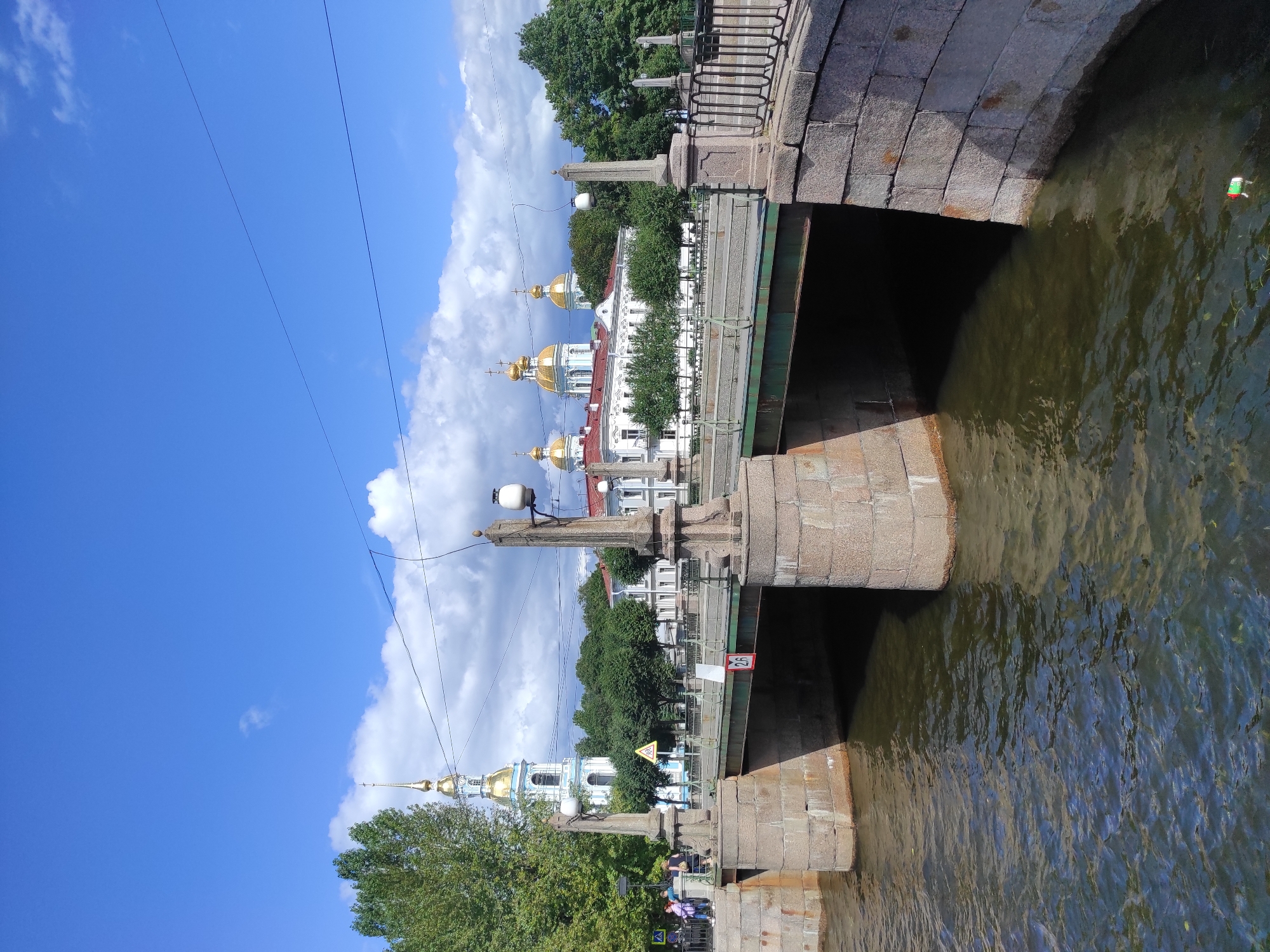 A wide shot depicts a canal in Saint Petersburg, Russia, on a sunny day with some clouds. A stone bridge with granite abutments spans the canal, supporting a road with a railing. On the bridge are streetlights atop granite posts. In the background, a white building with a gold-domed spire, a church, and other buildings are visible, surrounded by trees. Several people are standing on the left side of the bridge. The canal water is murky and reflects the sky and surroundings. A small piece of litter is floating in the water on the right side of the canal.