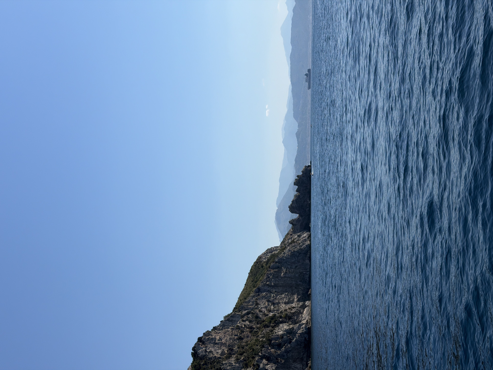 The image depicts a vertical split view. The right side shows a dark blue body of water, likely the sea, with visible surface texture and waves. The left side features a blue sky with a gradient from lighter to darker shades. In the middle ground, there's a rocky, mountainous terrain, partially covered with vegetation. The topography extends into the distance with silhouettes of distant hills or mountains. Based on the location, it is assumed the rocky terrain is part of the Amalfi Coast, specifically near Maiori, Italy. No people or other specific objects are discernible.