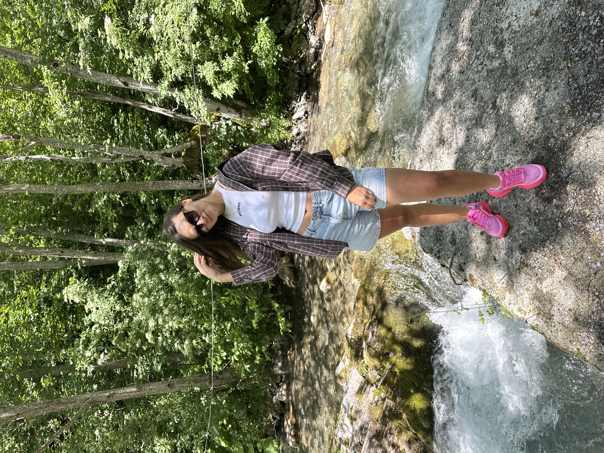 In Jabuka, Montenegro, a young woman stands on a rocky shore beside a flowing stream. She is wearing a white tank top with text, denim shorts, a plaid jacket, and bright pink sneakers. Her hand is raised, touching her hair, and she is looking towards the camera with sunglasses on. The background is filled with lush green trees, creating a natural, outdoor setting. The water in the stream is clear and appears to be moving rapidly, with small rapids visible. The sunlight filters through the trees, casting shadows on the ground.