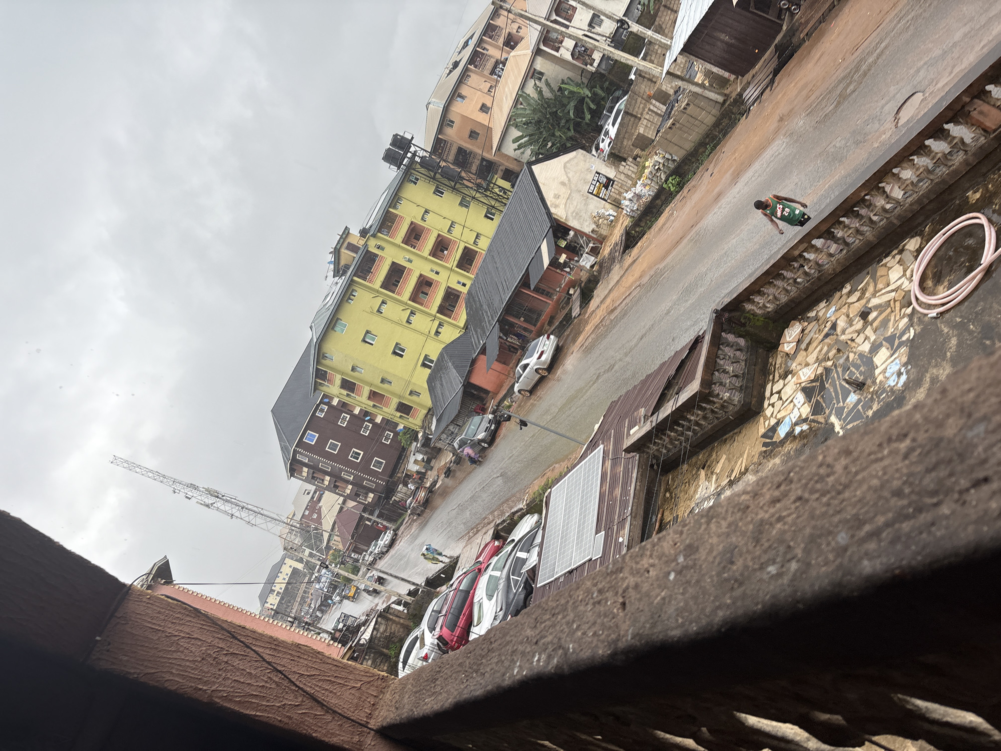 In Nkpor, Nigeria, on a rainy day, a person wearing a green and black sleeveless shirt with the number 20 walks across a wet street. The street is lined with parked cars, including a red SUV, a silver sedan, and a black sedan. Buildings with multiple stories and balconies are visible on either side of the street. One prominent building is painted yellow. A tall communication tower stands in the background. The scene is viewed from a higher vantage point, possibly a balcony, which shows a weathered railing and some solar panels on a rooftop in the foreground. Debris and a coiled pink hose are also present on the rooftop. The sky is overcast.