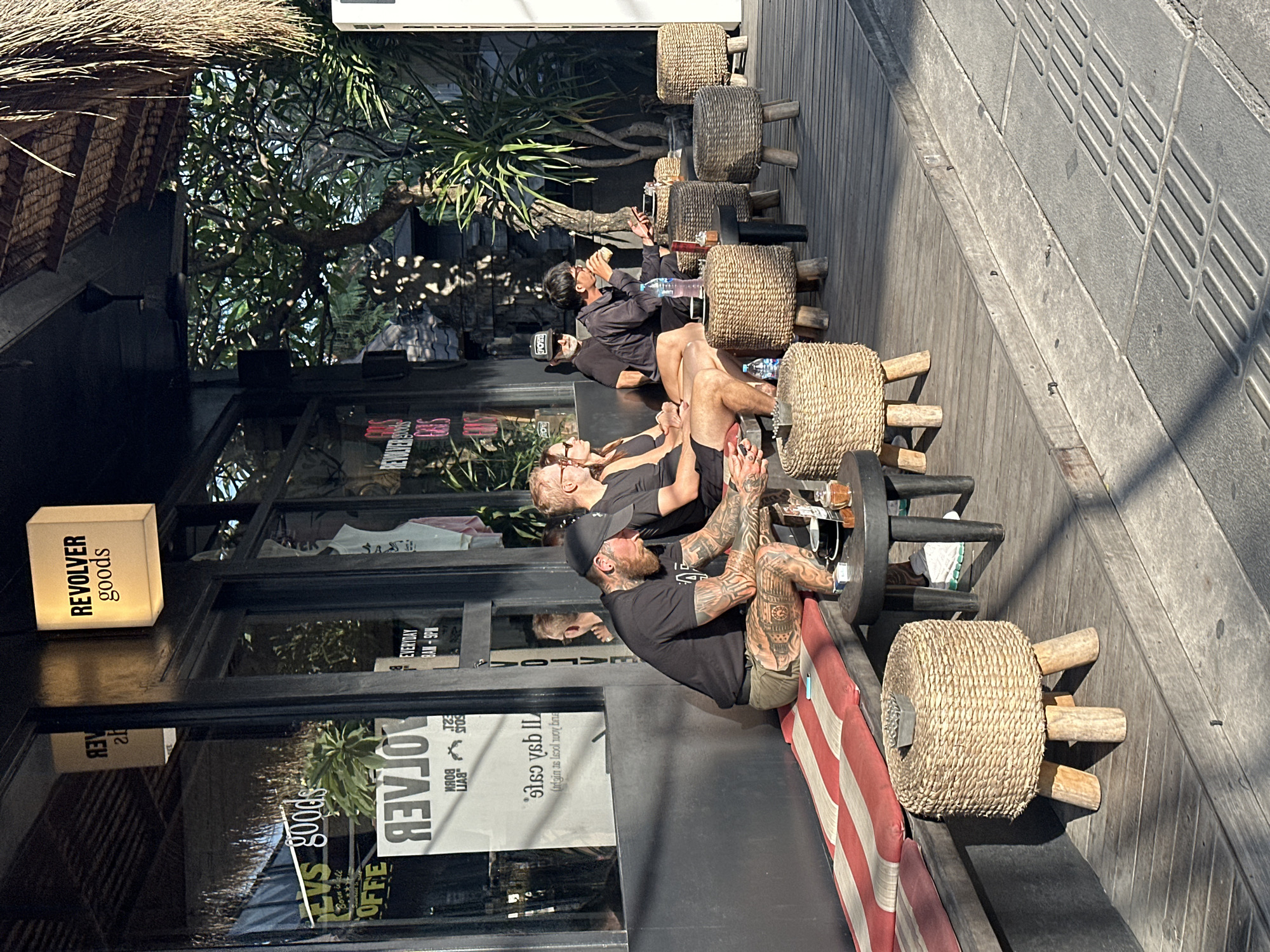 In North Kuta, Indonesia, outside a establishment with a thatched roof and a black facade, several individuals are seated on a wooden deck. To the left, a man with extensive tattoos on his arms and legs is wearing a black t-shirt and khaki shorts, sitting on a red and white striped bench. Next to him, a woman in a black tank top and shorts is seated, and a man in a black t-shirt and shorts is behind her. Further to the right, a man in a dark hoodie is holding a cup and a phone, and a man in a black t-shirt and glasses is seated. Several woven stools with wooden legs are placed around the seating area. A sign on the building reads "REVOLVER goods". The scene is illuminated by sunlight, casting shadows on the wooden deck and the street.