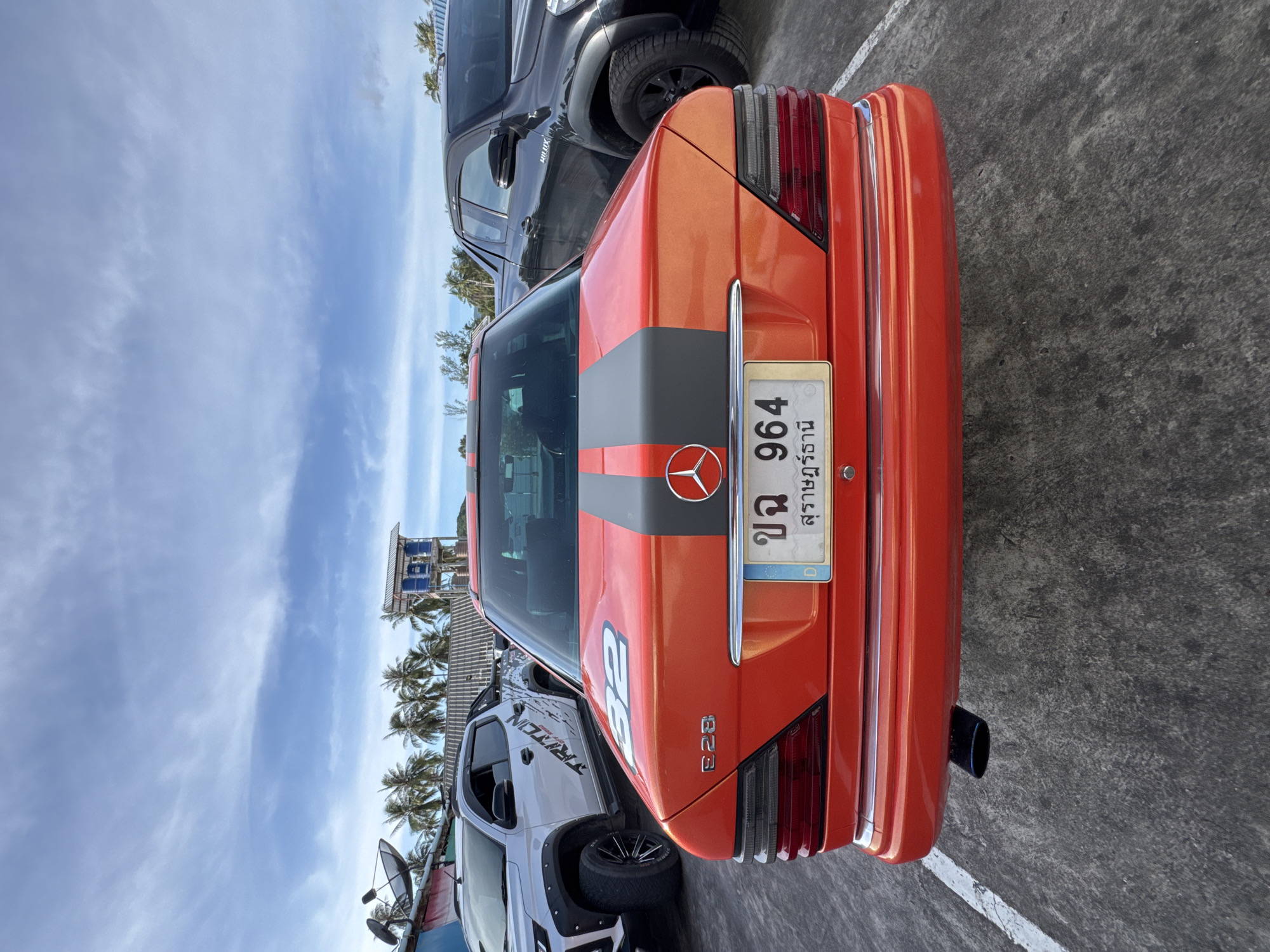 An orange Mercedes-Benz sedan with racing stripes and the number 32 on the trunk is parked on asphalt in ตำบลเกาะพะงัน, Thailand. The car has a blue and white license plate with Thai characters and the number 964. Behind the Mercedes, a black pickup truck is parked, and to the left, a white pickup truck with the word "TRITON" on its side is visible. In the background, palm trees and a small building with a blue roof and a water tower stand against a cloudy sky. The scene appears to be an outdoor parking area, possibly near a beach or resort, given the presence of palm trees and the clear sky.