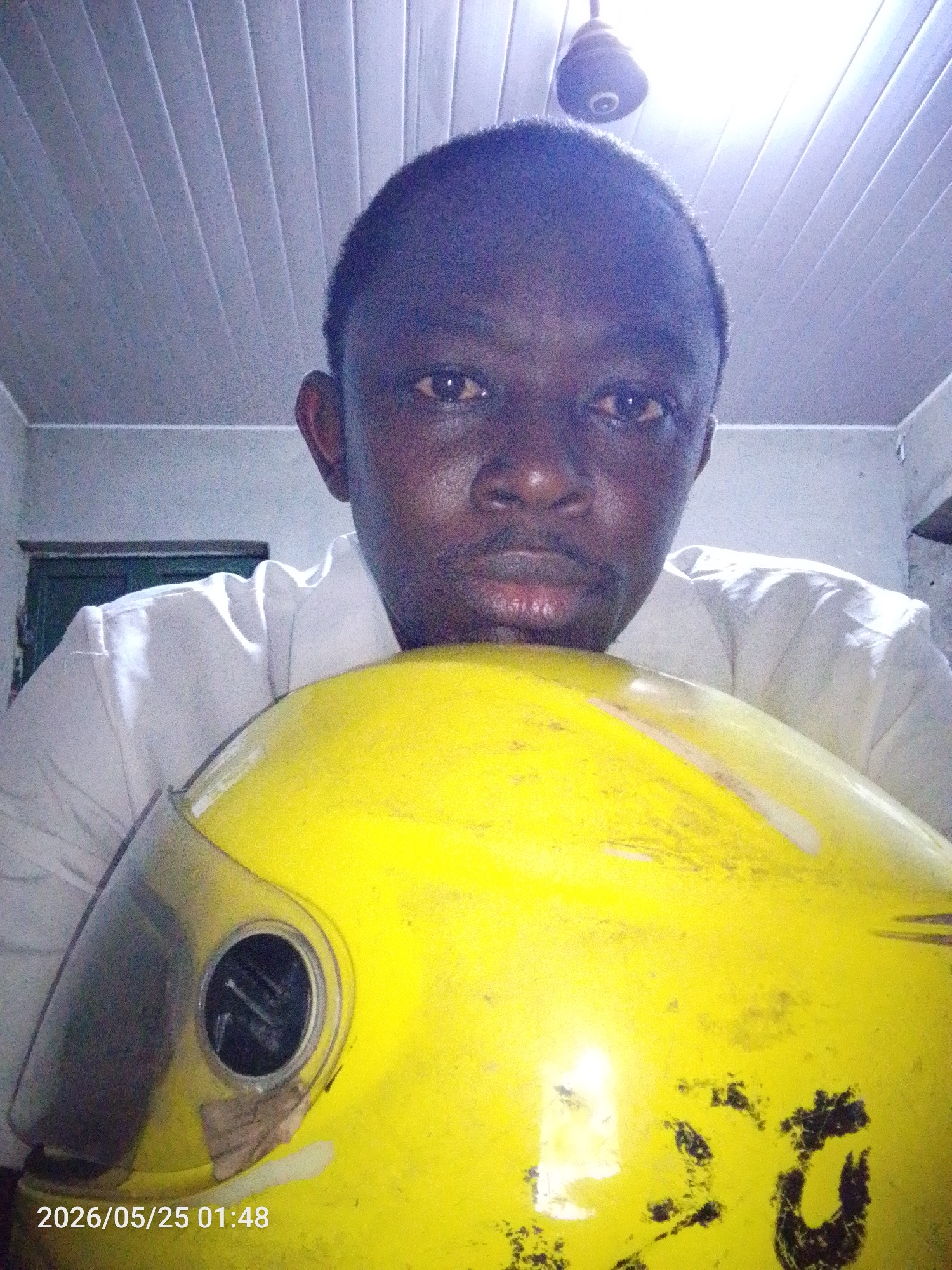A person is holding a yellow helmet. The person has dark skin, short hair, and is wearing a white shirt. The helmet appears used and has a partially obscured visor. The ceiling has paneling and a light fixture. The location is Lokoja, Nigeria (based on the likelihood of the image originating from there given the image content). The precise location within Lokoja is unknown. The image includes a timestamp of 2026/05/25 01:48.
