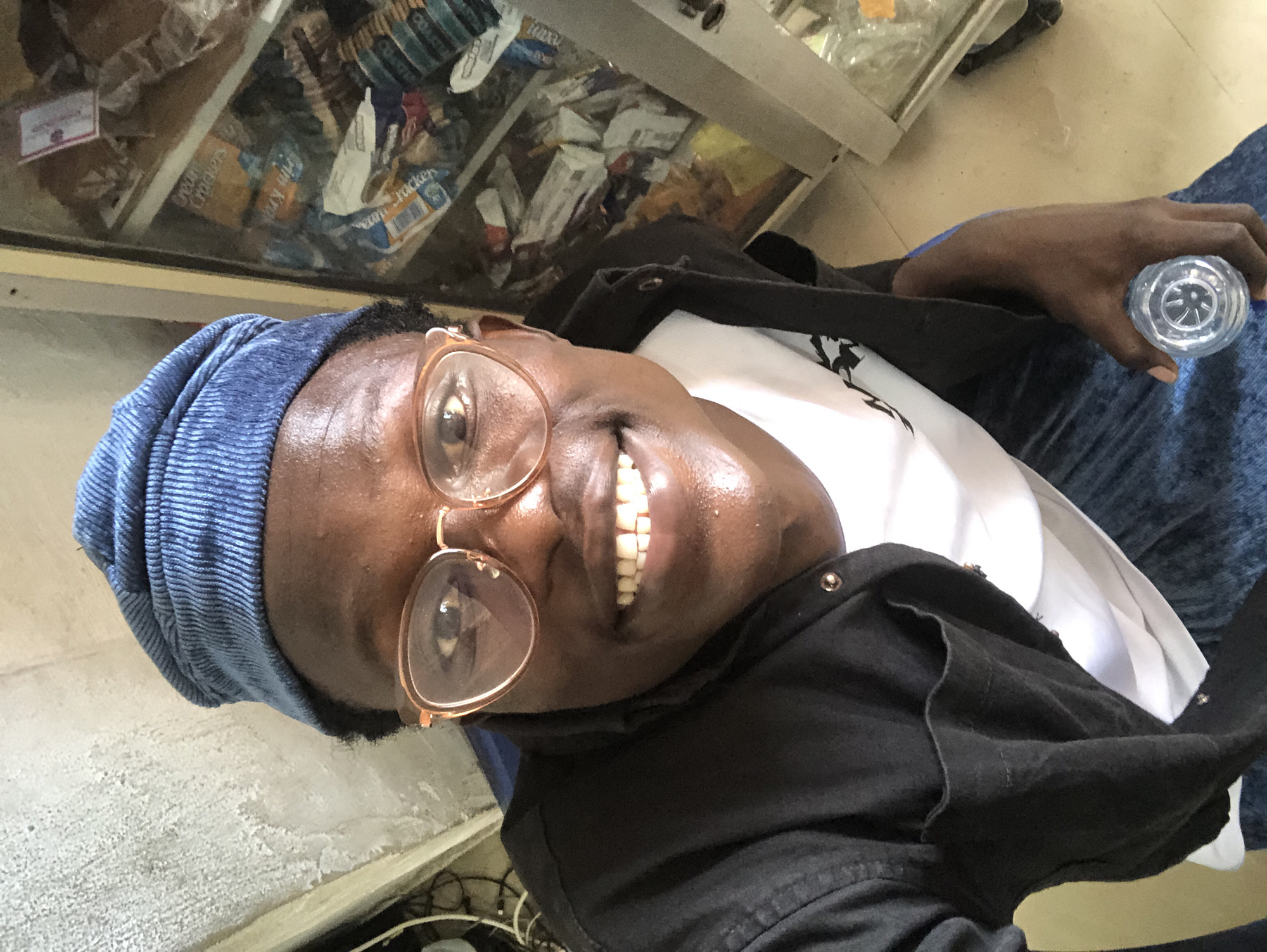 The image shows a close-up, high-angle selfie of a young Black man wearing a blue corduroy beanie, gold-rimmed glasses, a black unbuttoned shirt over a white t-shirt, and blue jeans. He is smiling broadly, showing his teeth. He is holding a clear plastic water bottle in his right hand. In the background, to the right, is a glass-fronted display cabinet filled with various packaged food items, possibly in a shop. To the left, the background appears to be a textured, light-colored wall with some wiring visible. The lighting suggests an indoor environment. The man's skin appears to be slightly sweaty. The overall scene is casual and personal. The mention of Ado, Nigeria, implies this location is a possible setting for the image.