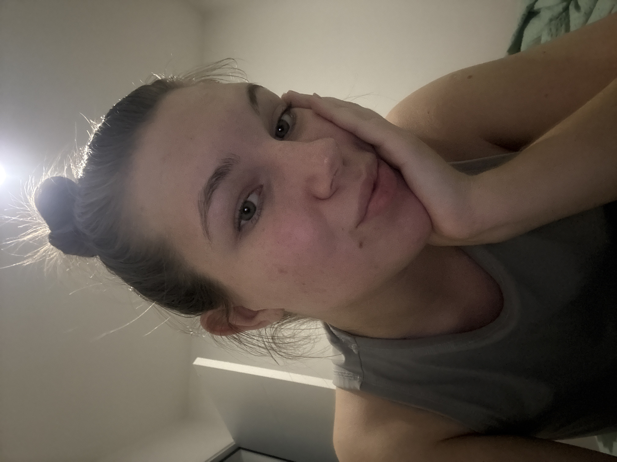 A woman is lying down, resting her face on her hand. She has her hair pulled back in a bun. She is wearing a gray tank top. The background appears to be an interior space, with a wall and a glimpse of what might be bedding. The location is not discernible from the image.