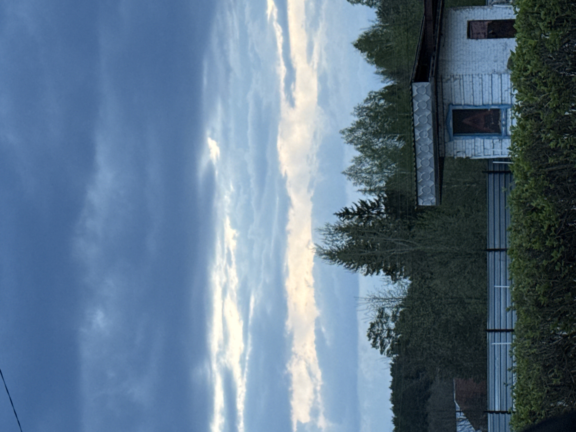 The image shows a vertically oriented view of an outdoor scene. The sky occupies the majority of the left side, featuring dark, cloudy formations interspersed with brighter, narrow bands of clouds. On the right side of the frame, a house is visible, with white siding and blue trim around windows and decorative elements. Adjacent to the house is a dense green hedge, obscuring some details of the scene. A fence runs along the lower right of the frame. Trees are visible behind the house and the hedge, indicating a wooded area. The location is potentially Chaikovsky, Russia.