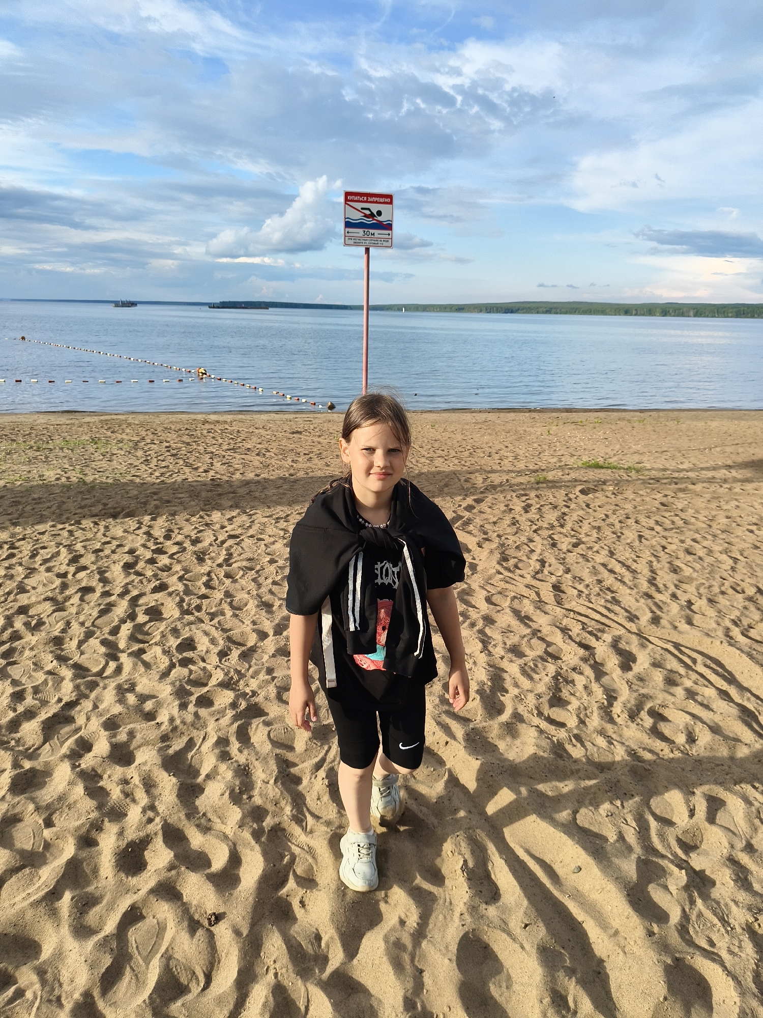 A young girl stands on a sandy beach facing the camera, with a calm body of water and a tree-lined horizon in the background. She is wearing a black hoodie draped over her shoulders, a black t-shirt with a graphic print, black shorts, and white sneakers. Her hair is pulled back. A red and white sign is visible behind her on a pole, depicting a swimmer and text in Russian. The sky above is partly cloudy with blue patches. The sand is textured with footprints and shadows cast from unseen objects. The scene appears to be at a public beach in the city of Chaikovsky, Russia, likely on the Kama River.
