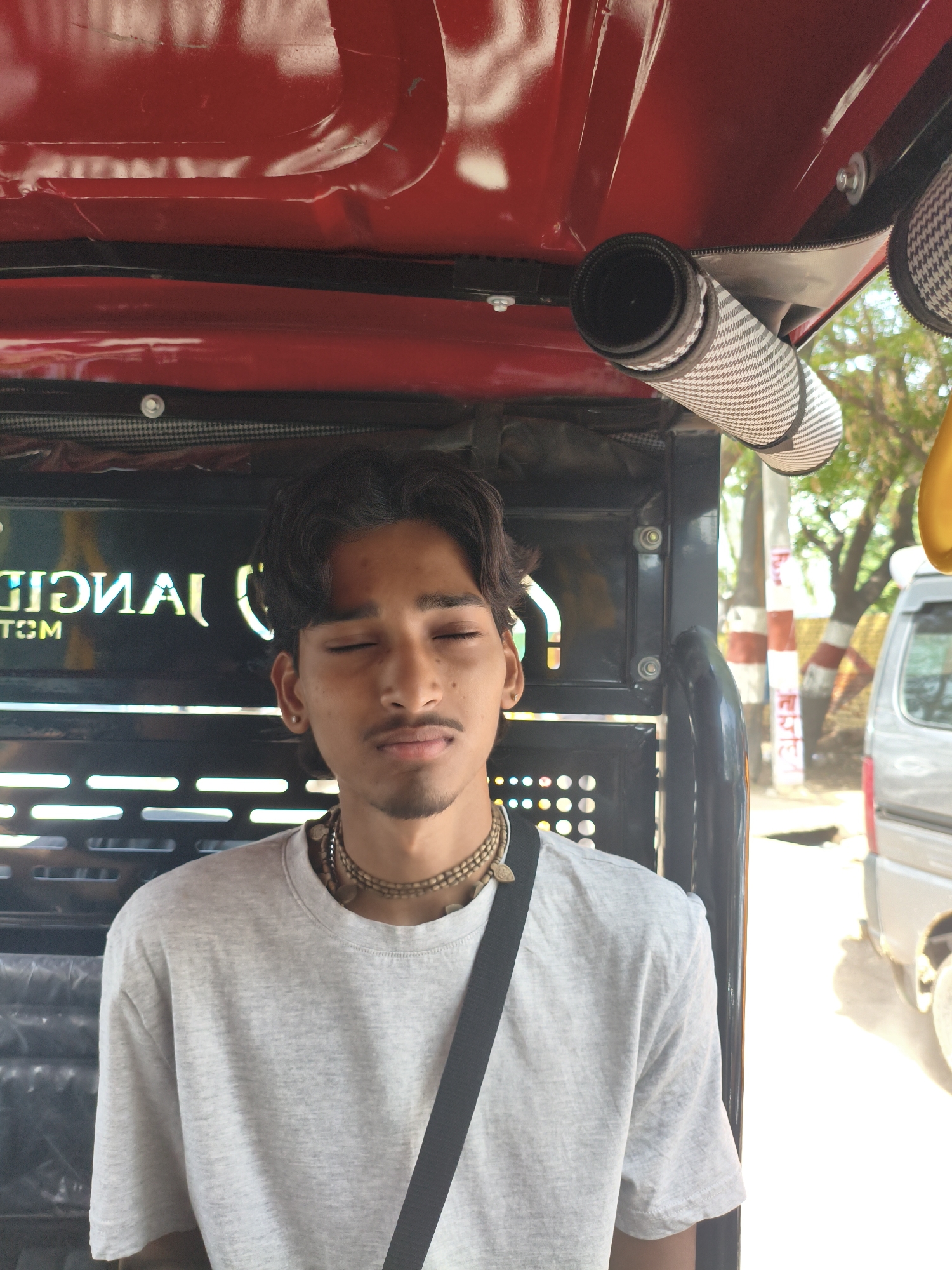 In Chhatikara, India, a young man with dark, wavy hair and a light mustache is seated inside what appears to be a motorized rickshaw. He is wearing a light grey t-shirt and multiple beaded necklaces. His eyes are closed, and his head is slightly tilted back. Behind him, the interior of the rickshaw features a black panel with white lettering that is partially obscured. The exterior of the rickshaw's roof is red. To the right of the rickshaw, trees and a white vehicle are visible, suggesting an outdoor, possibly urban or semi-urban, environment. A rolled-up patterned fabric is attached to the rickshaw's interior ceiling.