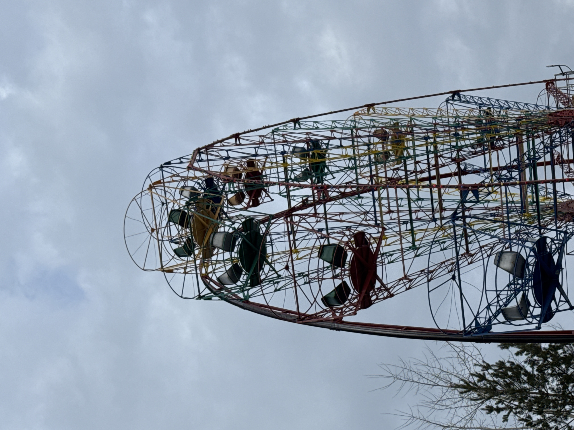 A low-angle shot captures a portion of a fairground ride under an overcast sky. The ride is constructed from a complex network of colorful metal scaffolding, supporting individual seating structures. The seats are mounted within circular frames, resembling the shape of a carousel. Trees are visible at the bottom right corner.