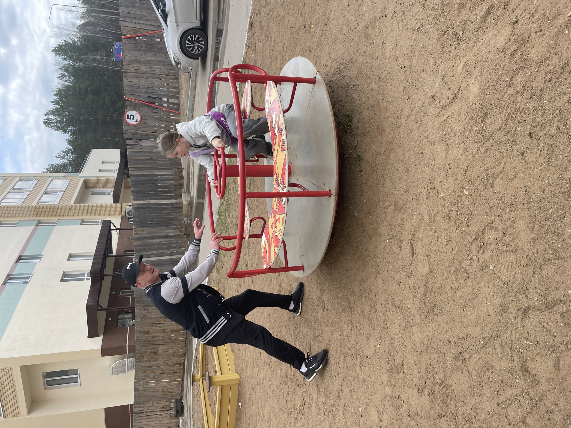 On a playground with a dirt surface in Chaikovsky, Russia, a man and a child interact with a merry-go-round. The child, wearing a light-colored jacket and a purple strap or bag across their chest, is leaning over the merry-go-round's edge. The man, dressed in a black jacket with white stripes and black pants, is standing beside the merry-go-round with his arms raised. Buildings, a wooden fence, and a parked car are visible in the background. A speed limit sign is present on the fence near some trees.