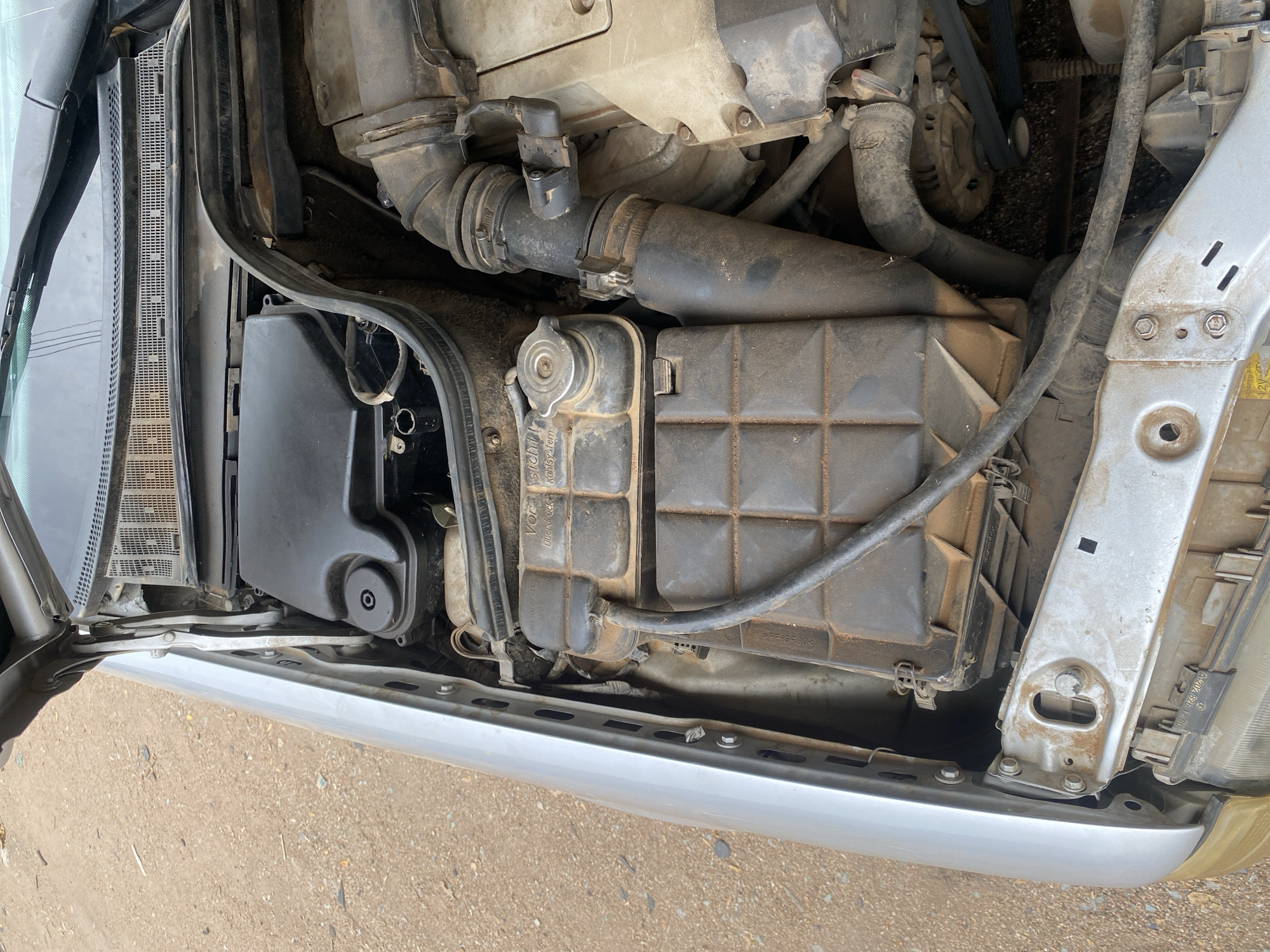The image shows an open car hood, revealing the engine bay of a silver sedan, likely parked outdoors on a dirt road in Jalingo, Nigeria. The engine components are covered in a thick layer of dust and dirt, suggesting it has been exposed to the elements. Key visible parts include the coolant reservoir, air intake system, and part of the engine block. The car appears to be older, with some visible wear and tear on the exposed metal parts. The background consists of a sandy, unpaved surface. No people are present in the image, and no actions are being performed.