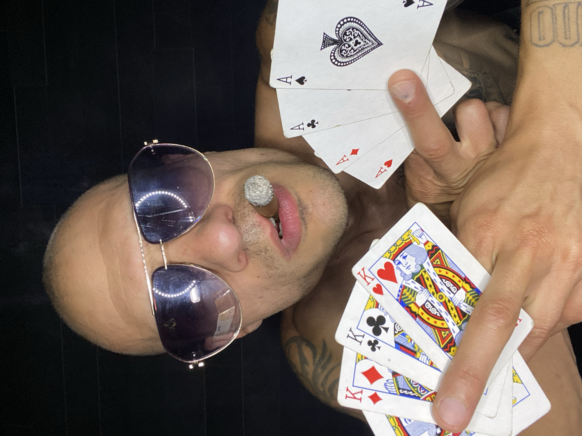 The image shows a man wearing sunglasses and holding playing cards and a cigar. He is looking at the camera with a cigar in his mouth. The playing cards consist of three kings and four aces. The man has tattoos on his left arm. The background is dark and appears to be a wall. There is no indication of the city of Pleven, Bulgaria, or any specific location within it.
