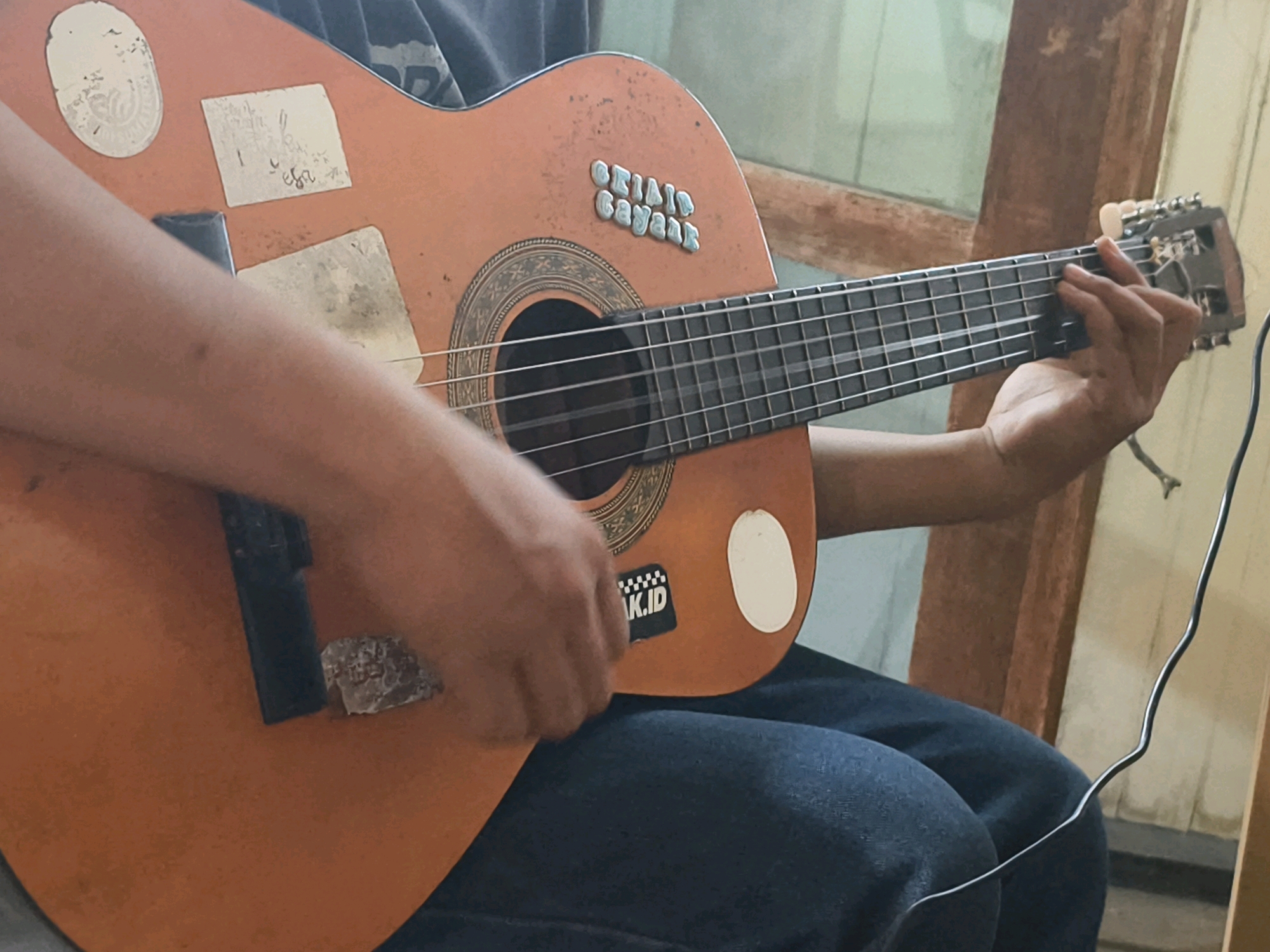 An individual is playing an acoustic guitar. The guitar is a light orange color and has several stickers on its body. The individual's hands are positioned on the fretboard and near the soundhole, strumming the strings. The scene appears to be indoors, with a wooden frame visible in the background, suggesting a rustic or traditional setting. The lighting is somewhat dim, casting soft shadows.