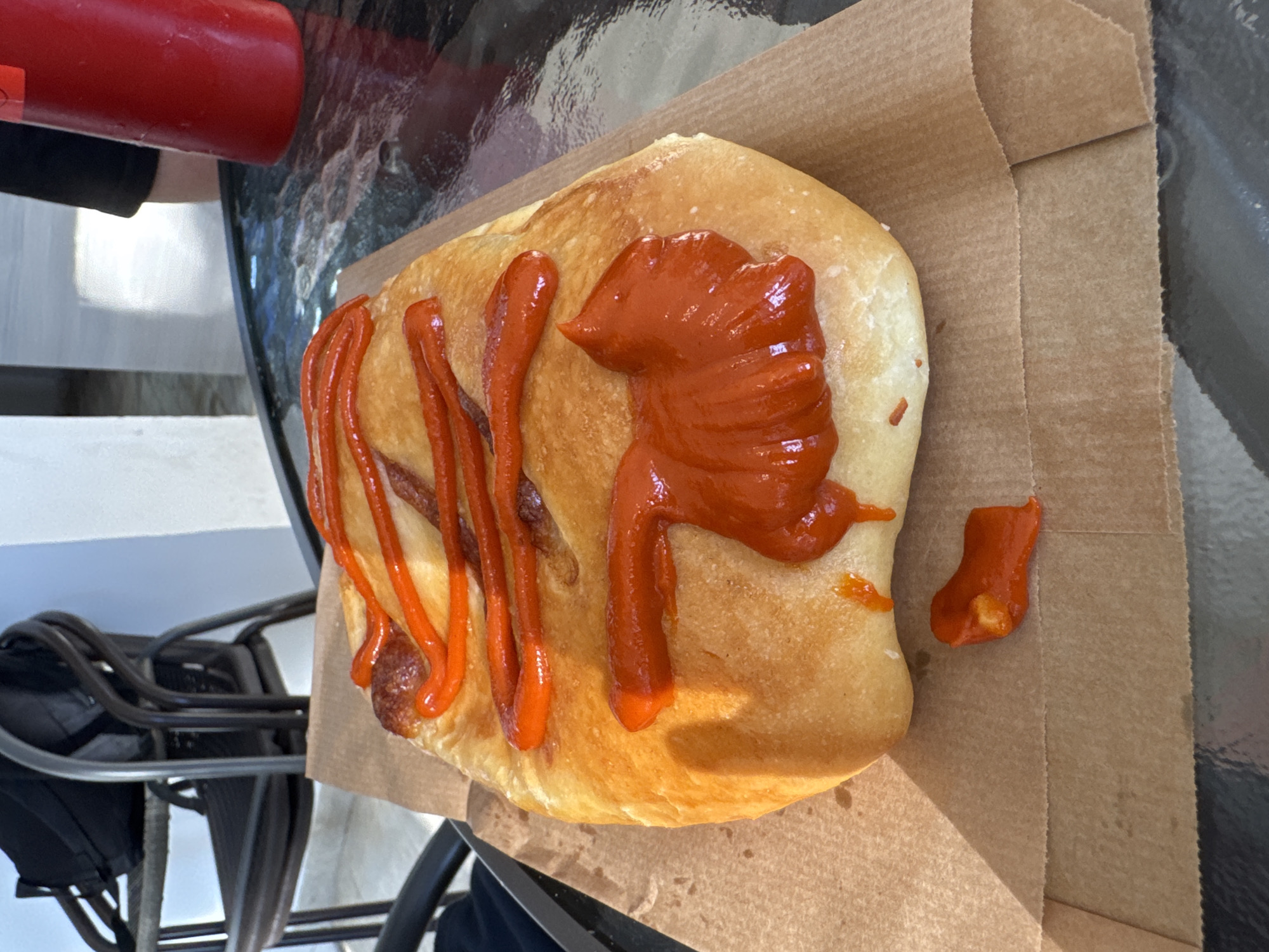A tan-colored sandwich sits on a brown paper bag atop a black metal table, likely outdoors due to the visible black metal chair and reflective table surface. The sandwich is topped with a generous amount of bright orange sauce, applied in drizzles and a large dollop on one side. A small piece of the sandwich with sauce sits separate from the main portion. A red bottle is visible in the upper left corner, positioned off the edge of the sandwich. The location is Unknown, Unknown.