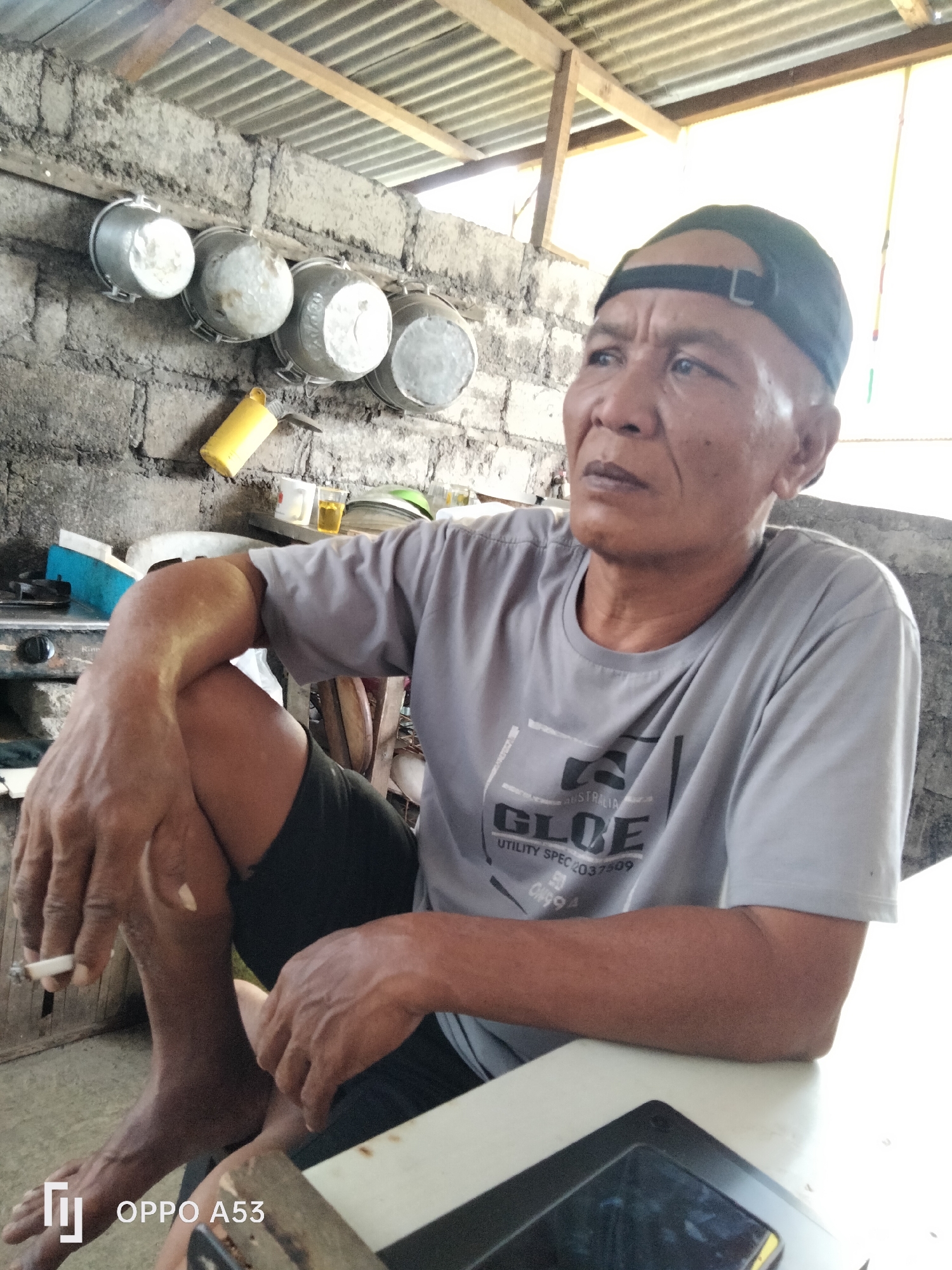 An overhead shot shows an adult male wearing a grey t-shirt and black shorts, sitting with his legs crossed while holding a lit cigarette in his left hand. He has a black baseball cap on his head and his gaze is directed upwards and to the right. Behind him, there are four metal pots hanging from a brick wall, and a yellow object hanging near them. The scene appears to be in a rustic outdoor setting, possibly a kitchen or dining area, with a corrugated metal roof overhead. The overall impression is one of a casual, everyday moment.