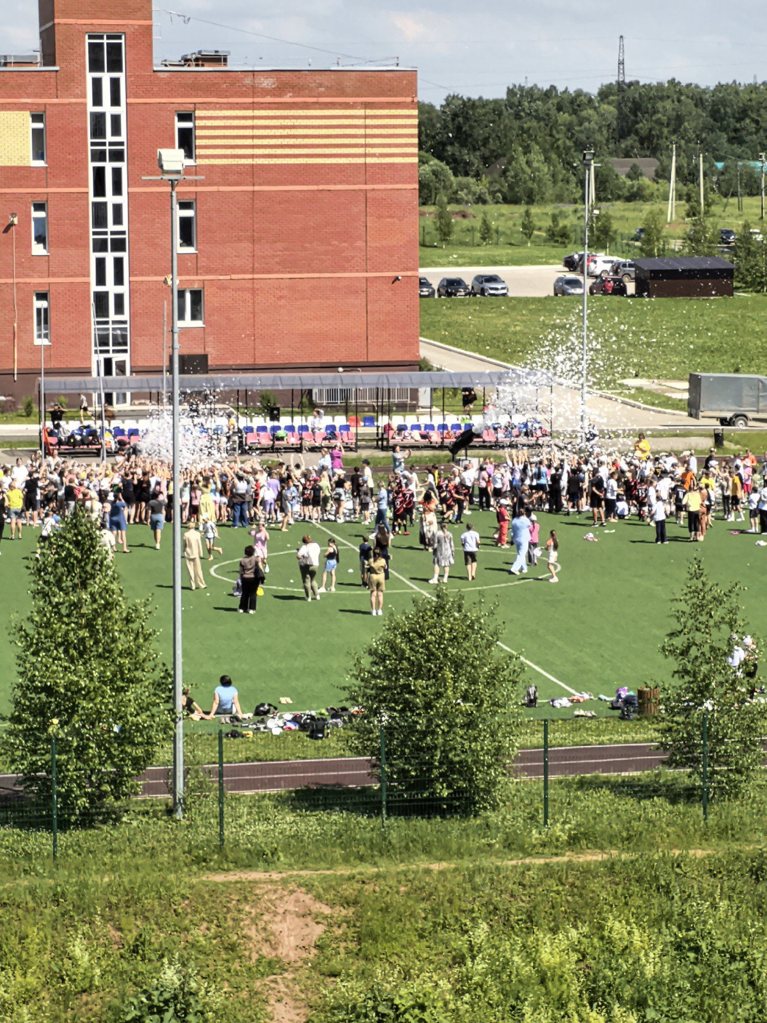 In Фролы, Russia, a large group of people, predominantly children and adults, are gathered on a bright green artificial turf field, possibly a sports complex or school grounds. A brick building with yellow and orange horizontal stripes is visible in the background, along with a covered seating area with blue and red benches. The group is dispersed across the field, with many people standing and interacting. A significant amount of white foam or bubbles is in the air, concentrated above a central area of the field where many individuals are looking up or reaching out. Several trees with dense green foliage are scattered across the field and its perimeter, and a chain-link fence runs along the foreground. Beyond the field, a parking lot with several cars, a dark brown storage shed, and more trees and vegetation are visible, with a power line tower in the distance. The sky is overcast with some visible sunlight, suggesting a daytime event.