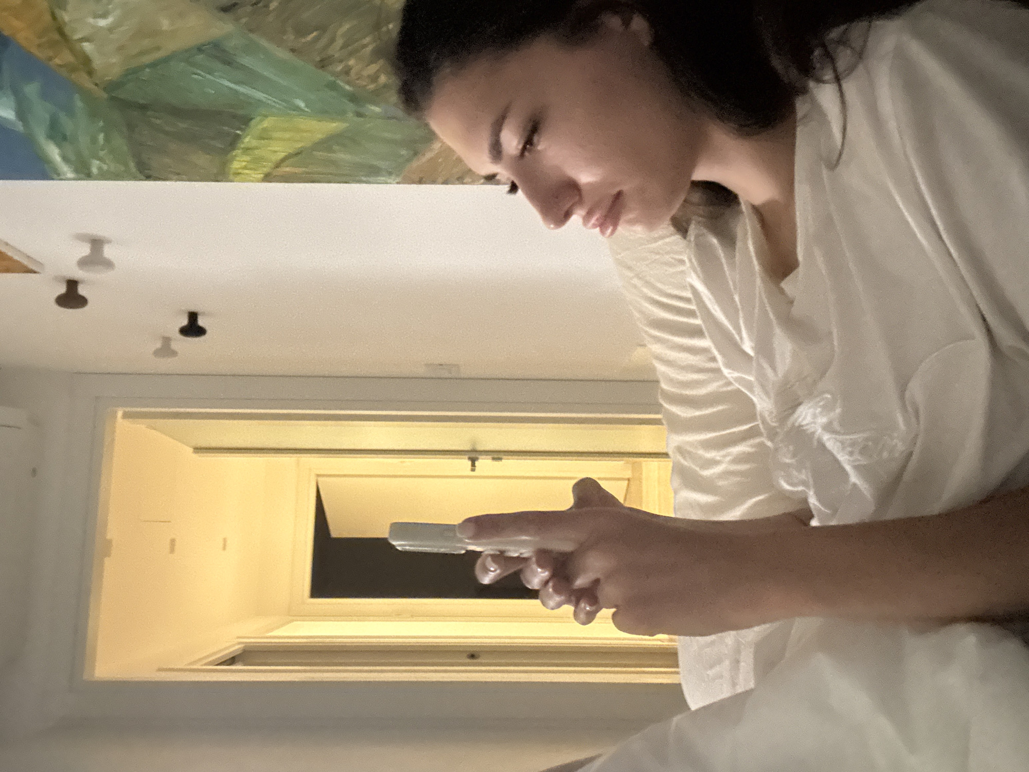 An overhead indoor shot shows a woman lying on a bed, propped up by pillows, holding a white remote control to her face. Her right arm is extended, with her fingers curled around the device, while her left arm rests on her chest. The woman is looking at the remote control with a slight frown on her face. She is wearing a white, loose-fitting top, and her dark hair is spread across her shoulders. In the background, a hallway is visible, with several doors and a painting on the wall. The lighting is dim, with a warm glow emanating from the hallway. The scene is suggestive of a hotel room in Cupra Marittima, Italy.
