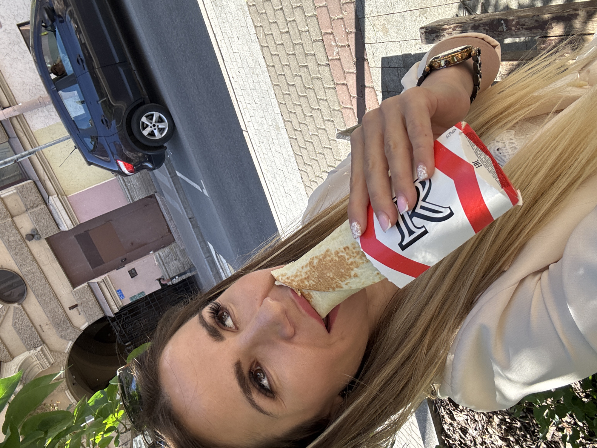 The image shows a woman taking a bite out of a burrito. She has long blonde hair and is wearing a light-colored jacket. She is also wearing a watch on her left wrist. The burrito is being held in a red and white wrapper. The woman appears to be outdoors, with a paved area and a building in the background. The sun is shining, indicating it is daytime. The scene is likely a street or outdoor seating area, possibly in Saint Petersburg, Russia, given the location context. The woman's expression is one of enjoyment as she eats her food.