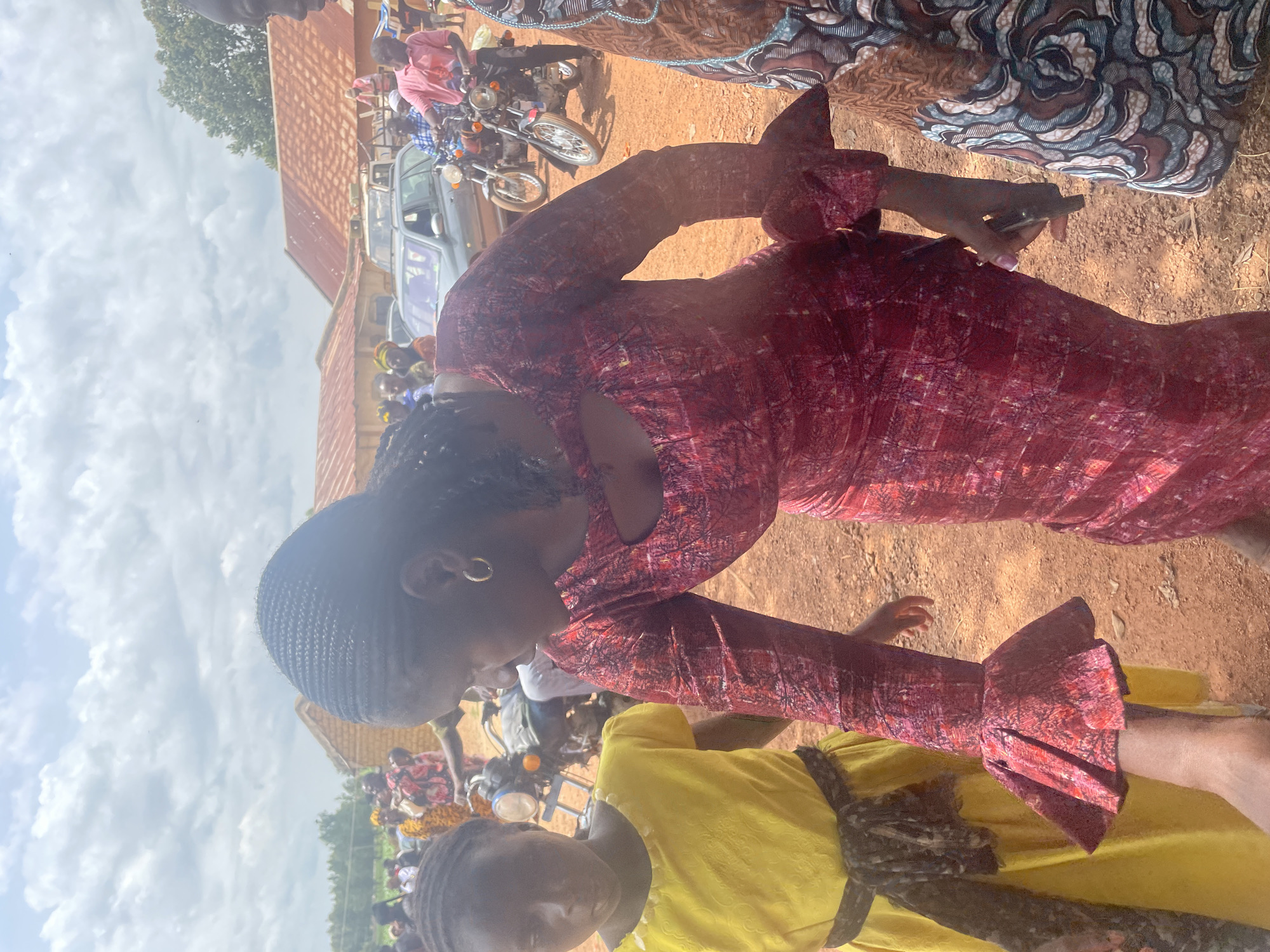 In an outdoor, dusty environment, likely in Unknown, Nigeria, a group of people are gathered. The focal point is a woman in a long-sleeved red patterned dress with flared cuffs, leaning forward and looking down. To her left, a young girl in a bright yellow dress stands, her face obscured. In the background, several motorcycles and a car are visible, suggesting a gathering or event. The buildings in the background appear to be simple structures with corrugated metal roofs. The sky is partly cloudy.