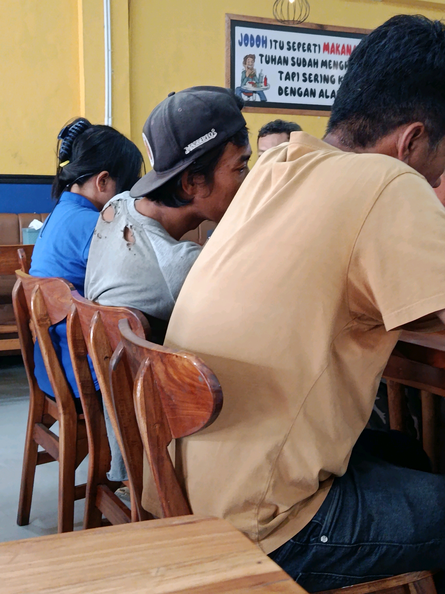 The image captures a scene at a restaurant in Bekasi, Indonesia, where three individuals are seated at a table. In the foreground, a person wearing a light brown t-shirt and dark jeans is partially visible from the back, facing away from the camera. To their left, another individual with a dark baseball cap worn backwards is seated, wearing a light grey t-shirt with several visible holes. Further to the left, a person in a bright blue shirt is also seated. Behind the seated individuals, a sign with Indonesian text is mounted on a yellow wall. The chairs are made of polished wood with an ornate design. The overall environment suggests a casual dining setting.