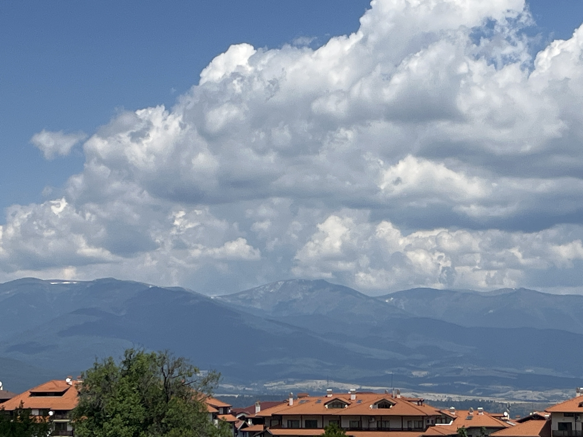 This is a medium-shot, eye-level, outdoor photograph of a mountainous landscape in Bansko, Bulgaria. The sky is a vibrant blue with a large formation of fluffy white and grey clouds dominating the upper half of the frame. Below the clouds, a range of blue-hued mountains stretches across the horizon, with some patches of snow visible on their peaks. In the foreground, the terracotta-tiled roofs of several buildings are visible, along with the lush green foliage of a tree. The buildings appear to be part of a residential or resort area. The overall scene depicts a clear, sunny day with a dramatic sky and a majestic mountain backdrop.