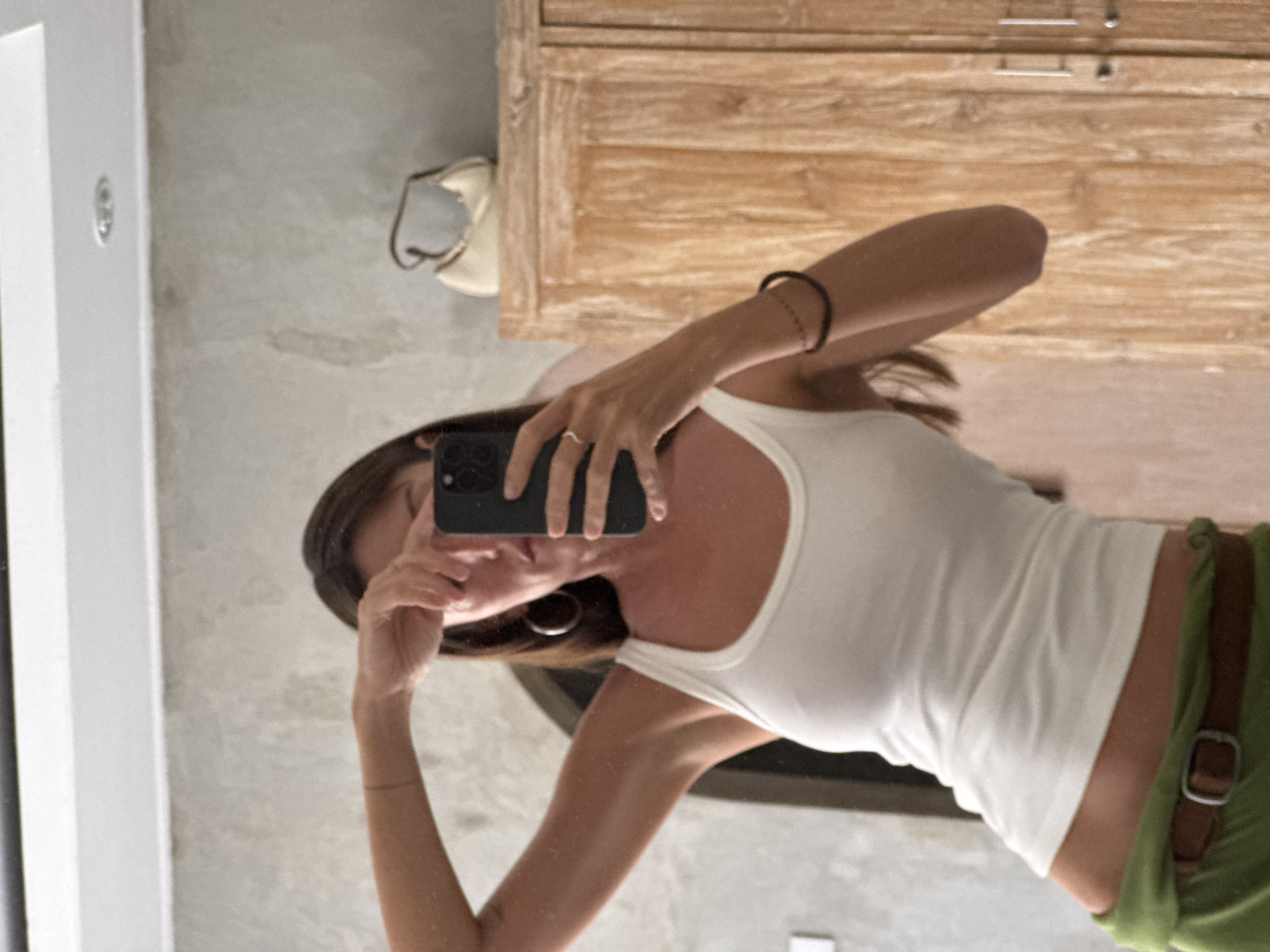 In North Kuta, Indonesia, a woman takes a mirror selfie in what appears to be a bathroom or dressing area. She is wearing a white tank top and green pants, cinched with a brown belt. A ring is visible on her left ring finger, and she has hoop earrings. Her right arm is raised, obscuring part of her face as she holds her phone. In the background, a light-colored handbag rests atop a wooden cabinet. The walls have a textured, concrete-like finish.