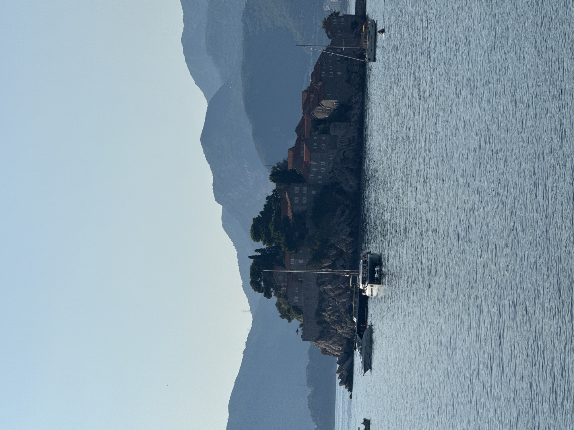 This is a landscape photograph featuring a scenic view of Sveti Stefan, a coastal town in Montenegro. The image is oriented vertically. In the foreground, the calm, blue water of the Adriatic Sea gently laps against a rocky island. Moored in the water are several boats, including a sailboat and a larger yacht. The island itself is densely vegetated, with trees and shrubs clinging to the rocky terrain.
Dominating the island are stone buildings with red-tiled roofs, indicative of traditional Mediterranean architecture. These structures are nestled into the hillside, creating a picturesque and historic settlement. Behind the island, a series of majestic, layered mountains rise in the background, their slopes covered in a hazy, bluish-green hue. The clear, light blue sky suggests fair weather and likely indicates it's daytime, possibly late afternoon or early morning due to the soft lighting.
The overall impression is one of tranquility and natural beauty, with the man-made structures harmoniously integrated into the stunning coastal landscape. There are no visible people or text in the image.