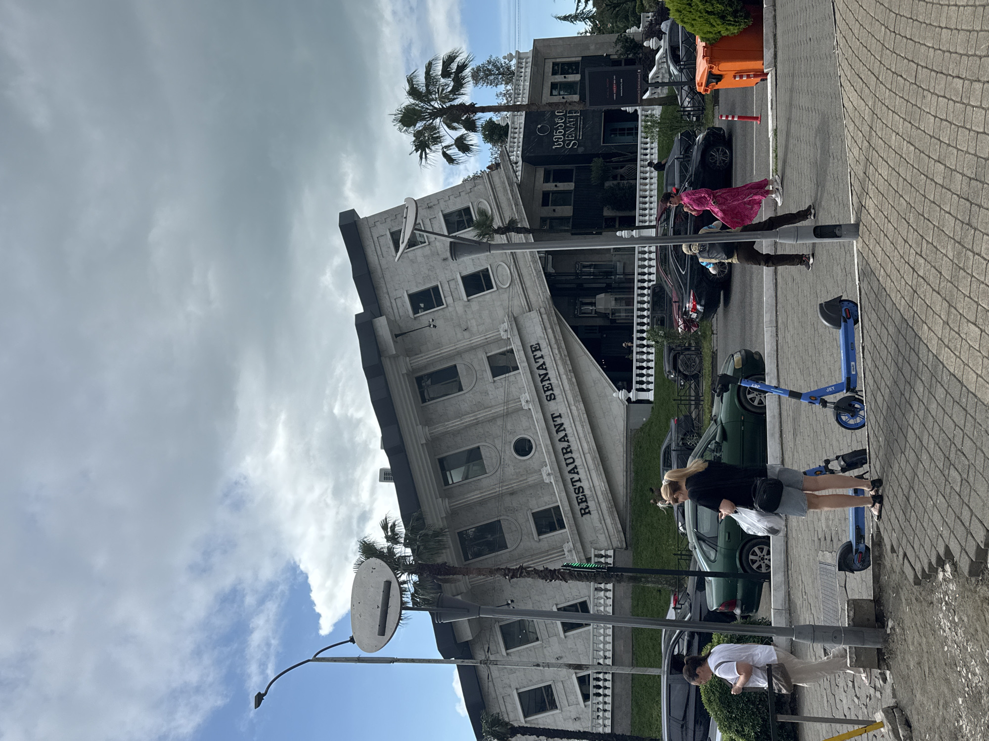 The image depicts a street scene in Batumi, Georgia, with an optical illusion building that appears to be tilted. The building has "RESTAURANT SENATE" written on its facade. Several people are present, walking on the sidewalk or standing near parked cars. One woman in a pink dress is walking away from the camera, while another woman in a black top and grey shorts is holding shopping bags. A third person in a white t-shirt is looking down. The scene appears to be set during the daytime, with a mix of sun and clouds in the sky. The street is paved with cobblestones, and there are palm trees visible, contributing to the tropical feel of the location. Electric scooters are parked along the sidewalk, suggesting a modern urban environment. The overall atmosphere is casual and everyday.