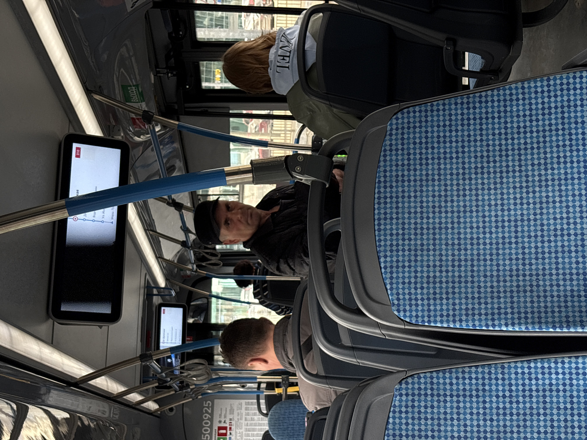 The image captures the interior of a public bus, likely in Khimki, Russia, based on the signage present. Several passengers occupy the bus, with a man in a black cap looking directly at the camera in the middle. In front of him, there is a seat, and behind him, there is a person wearing a jacket. Other individuals are present, seated further back in the bus. Informational displays are mounted on the interior of the bus, showing route information, and the seats are upholstered with a distinctive blue patterned fabric. Metal support bars with blue sections run along the roof of the bus, providing handholds for standing passengers.