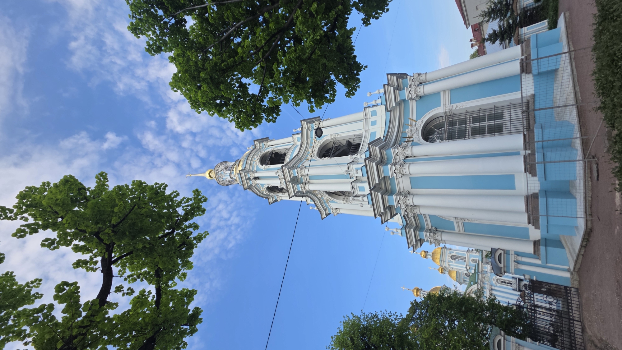 The image shows the exterior of the Saint Nicholas Naval Cathedral in Saint Petersburg, Russia, viewed from a low angle. The sky is blue with scattered clouds, and trees with green leaves frame the left side of the cathedral. The cathedral is white and blue with gold domes. Part of the building is under construction with metal fencing present.