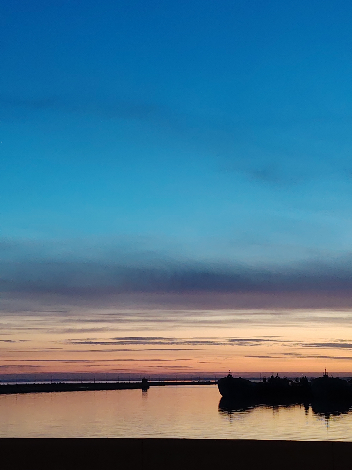 The image depicts a twilight scene in Chaikovsky, Russia. The sky transitions from a deep blue at the top to horizontal bands of purple, orange, and gold near the horizon. Wispy clouds are visible in the lower portion of the sky. Below the horizon line, a dark, still body of water reflects the colors of the sky. A breakwater stretches across the water's surface, with silhouettes of structures and poles visible on the breakwater. In the foreground, a dark silhouette of what appears to be a large barge or ship is anchored in the water. A dark, undefined landmass or structure is at the very bottom of the frame.