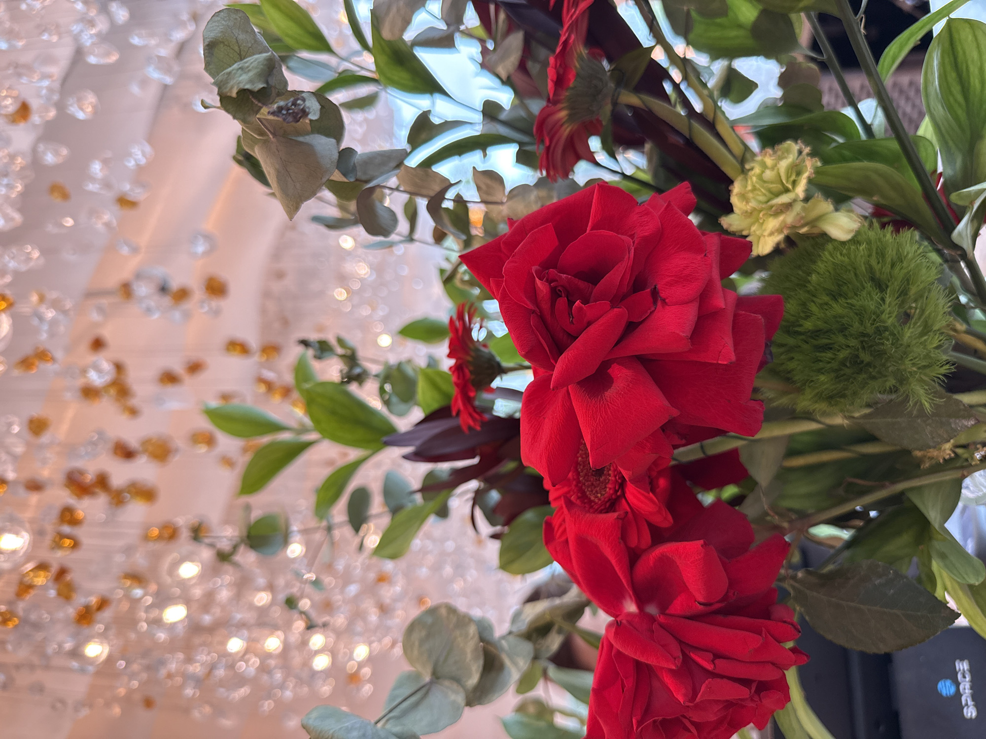 The image shows a close-up view of a floral arrangement featuring several vibrant red roses, green foliage, and some smaller red and pale yellow flowers. The background is softly blurred and illuminated with bokeh lights, suggesting an indoor setting with decorative lighting, possibly from a chandelier or string lights. The lighting in the background is a mix of warm amber and cool white, creating a festive or elegant atmosphere. No specific location in Nizhny Novgorod, Russia, can be identified from the image. The arrangement appears to be artfully composed, with the red roses being the dominant subject.