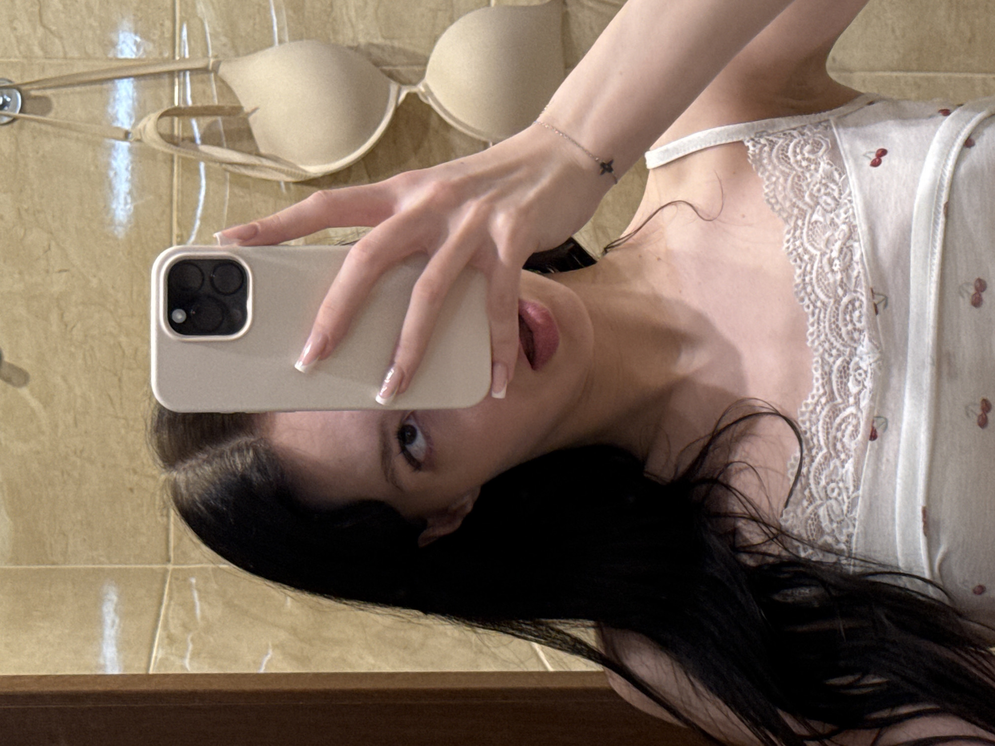 A young woman with long dark hair is taking a mirror selfie in a bathroom. She is wearing a white camisole with a lace trim and a cherry pattern. Her right hand is holding a white smartphone up to her face, obscuring part of it. A beige bra is hanging on the wall behind her. The walls appear to be tiled with beige tiles. The scene is consistent with a bathroom in an apartment in Khimki, Russia.